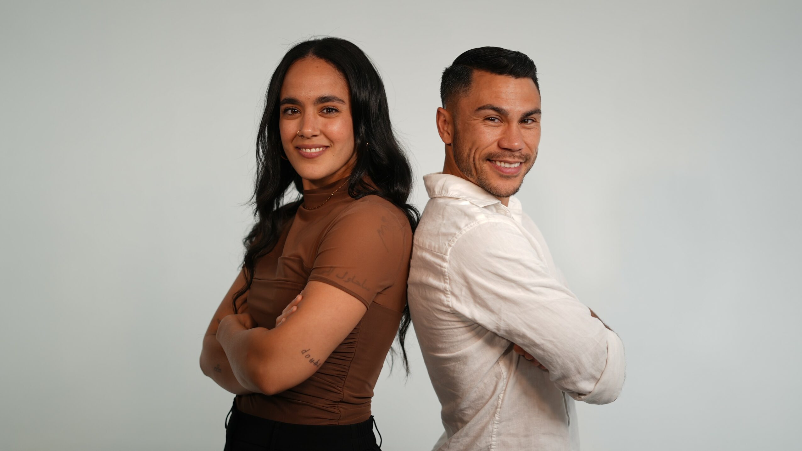 Two people standing back-to-back, smiling, with arms crossed, against a plain light background.