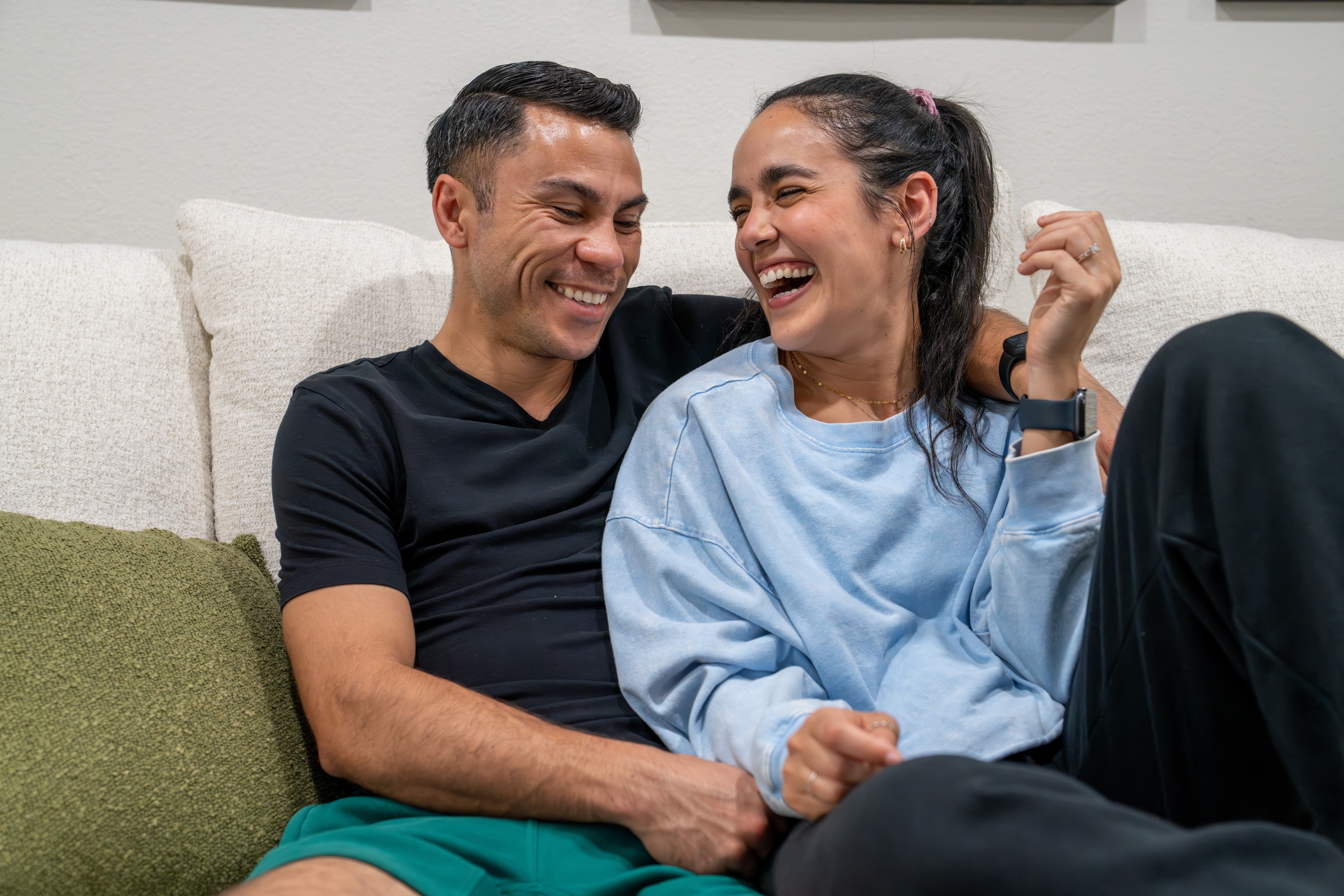 A man and woman sit on a sofa, smiling and laughing together, in a cozy indoor setting.