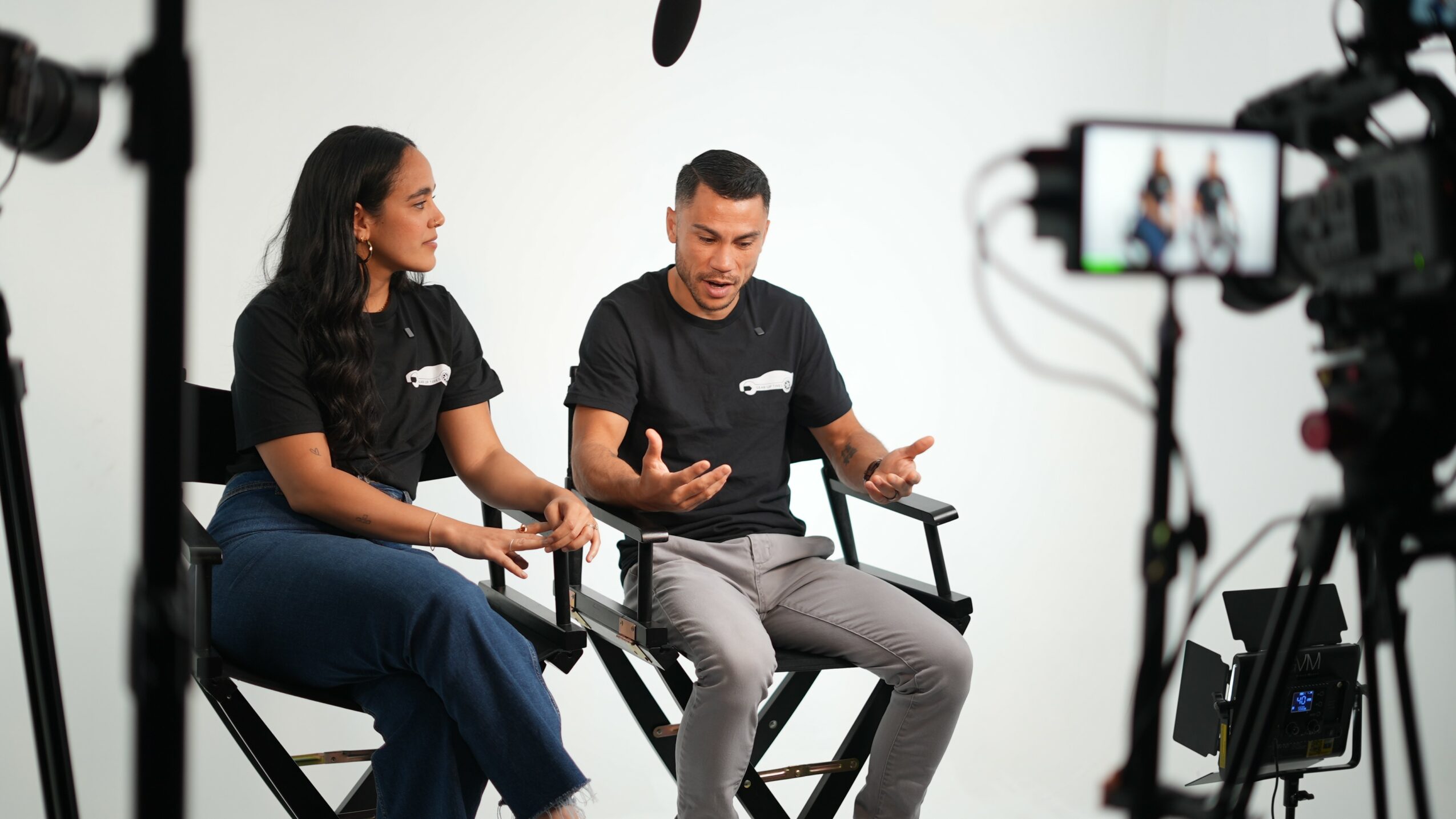 Two people sitting on chairs in a studio, being recorded with cameras and microphones, talking to each other.