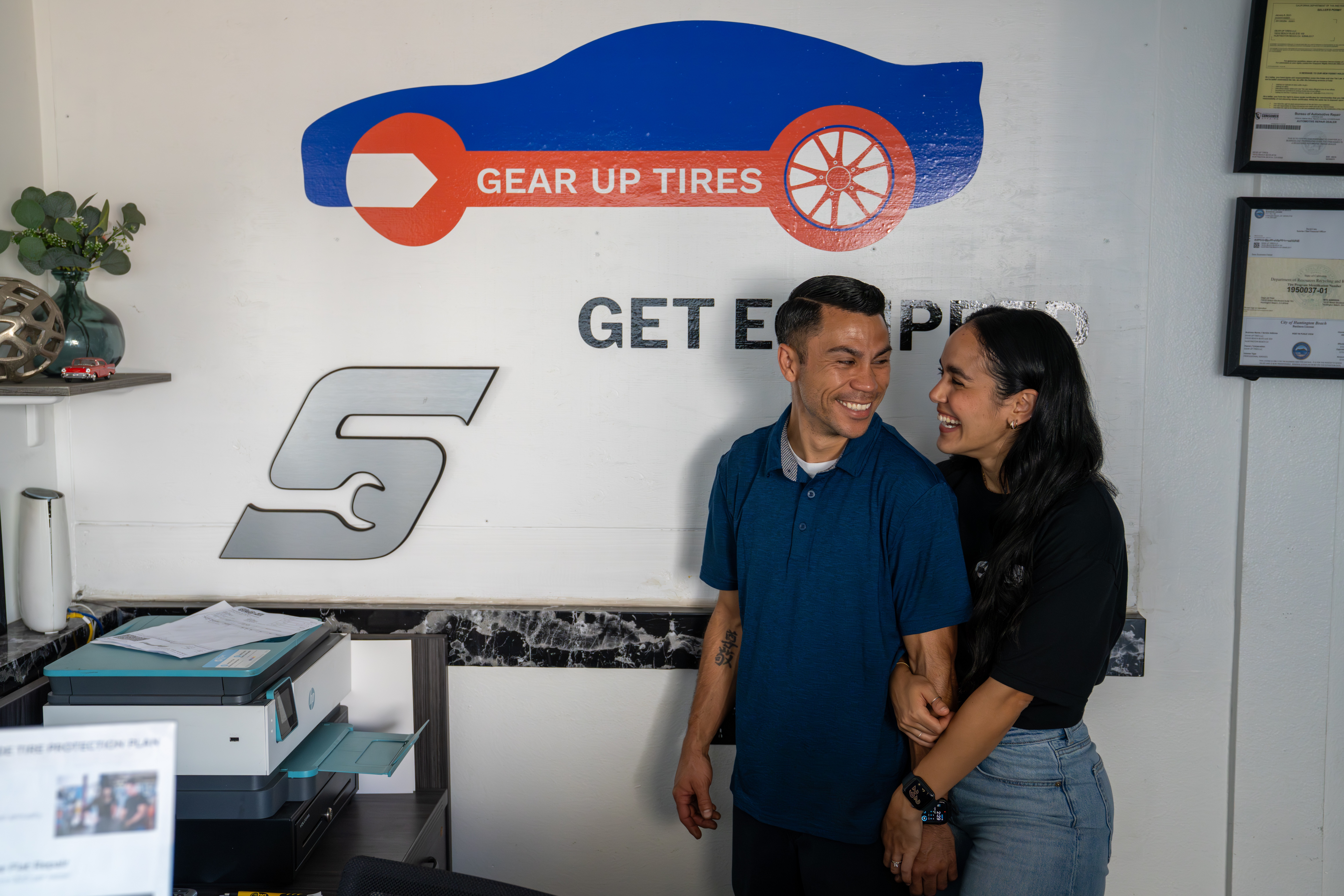 Two people smiling and talking in front of a wall with a logo and text, one person has a blue shirt, the other black.