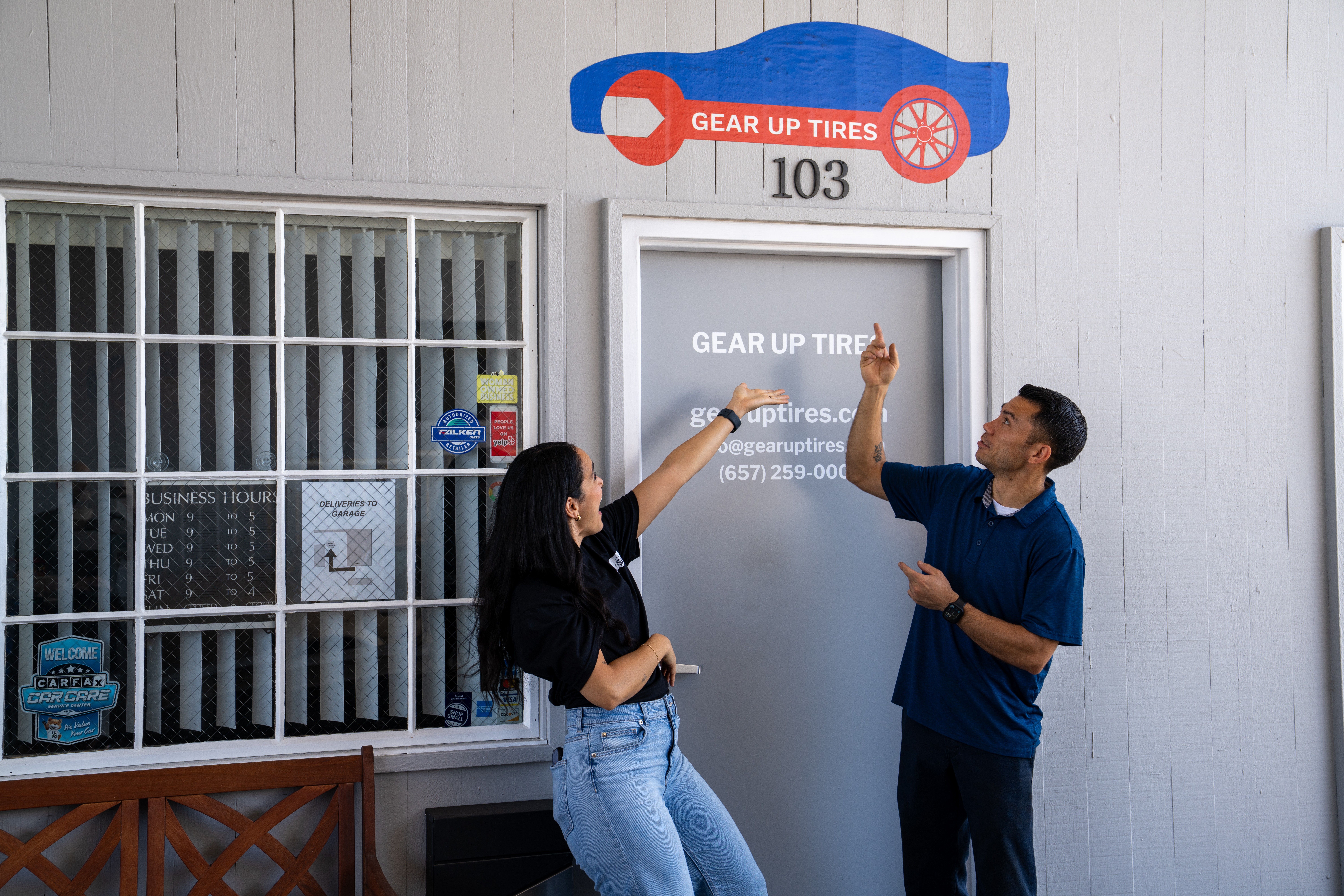 Two people standing outside a white building, one pointing at the door, the other holding a phone, near a window with bars.