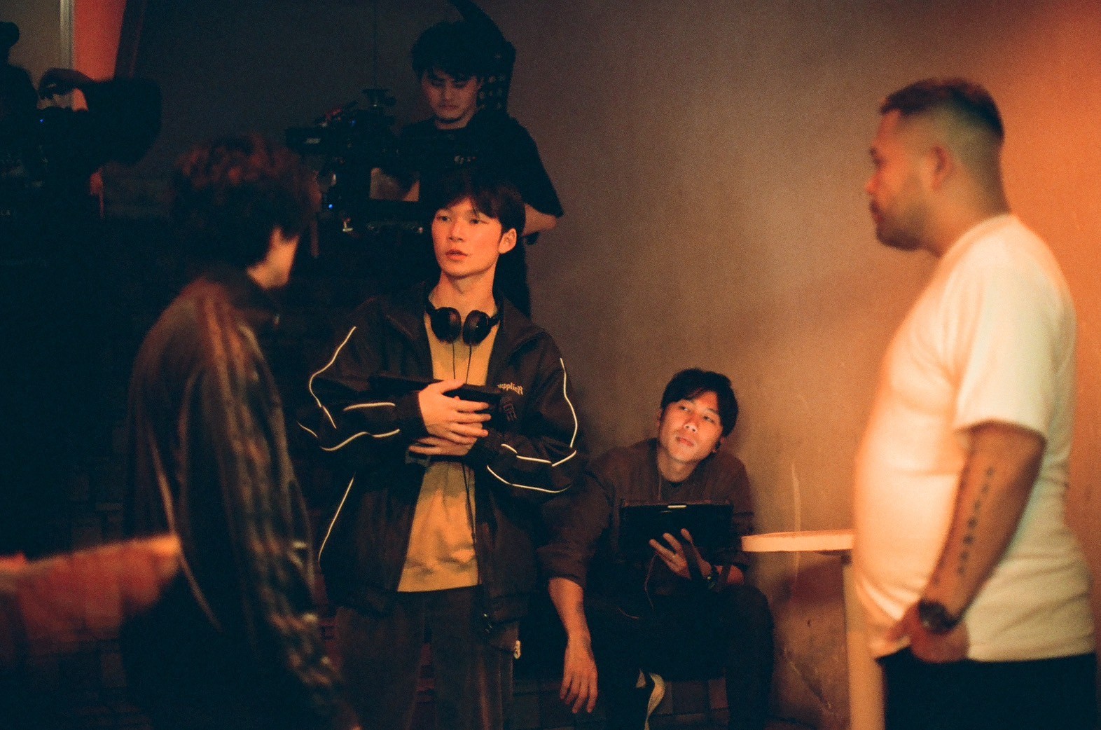 Group of young people in a dimly lit room, some standing and one sitting, engaged in conversation.