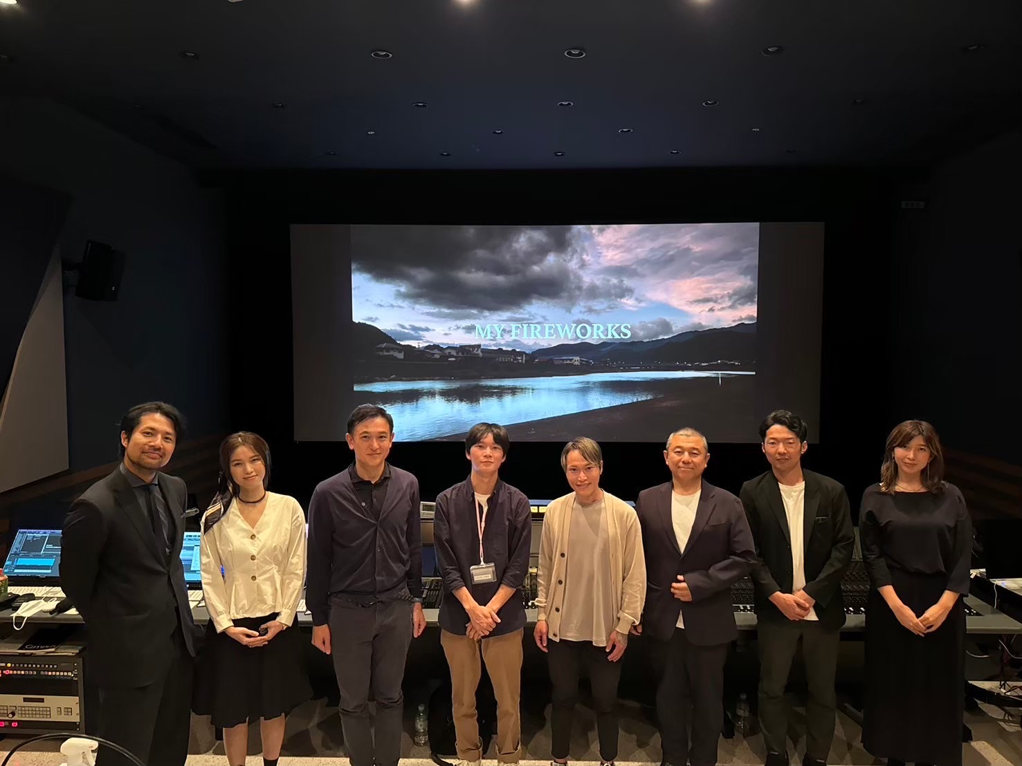 Group of seven people standing in front of a large screen displaying a landscape with water and clouds.