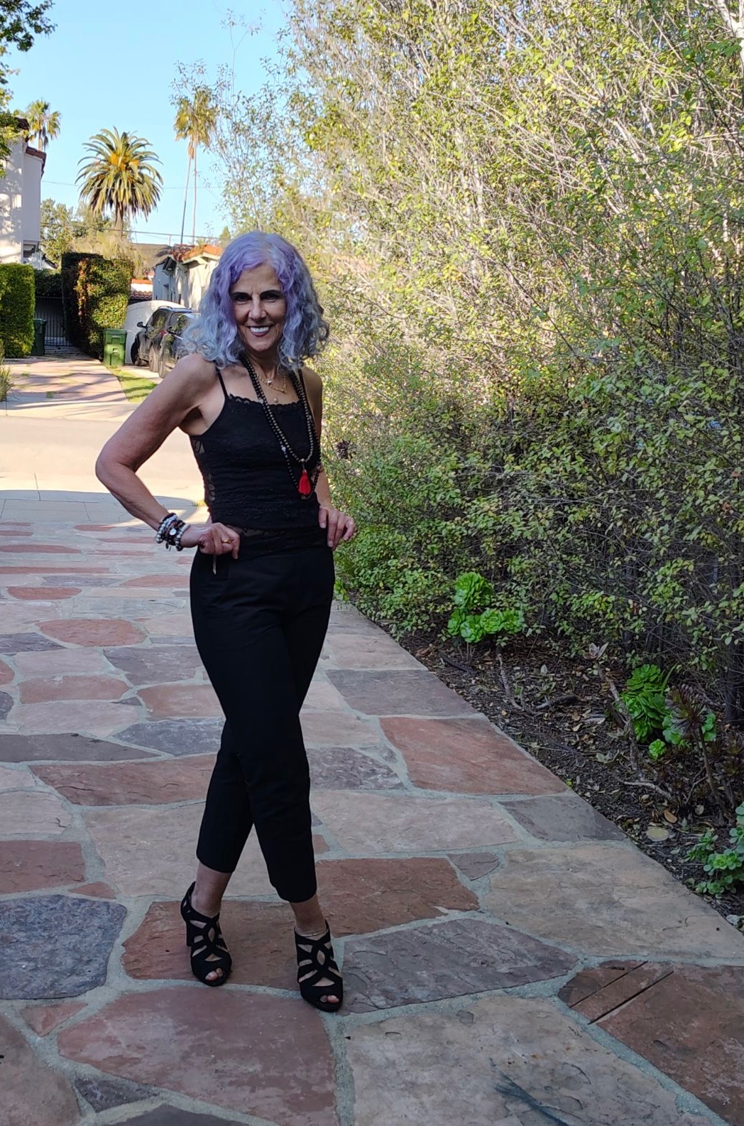 Woman with curly gray hair standing on a paved path outdoors, smiling, surrounded by trees and greenery.