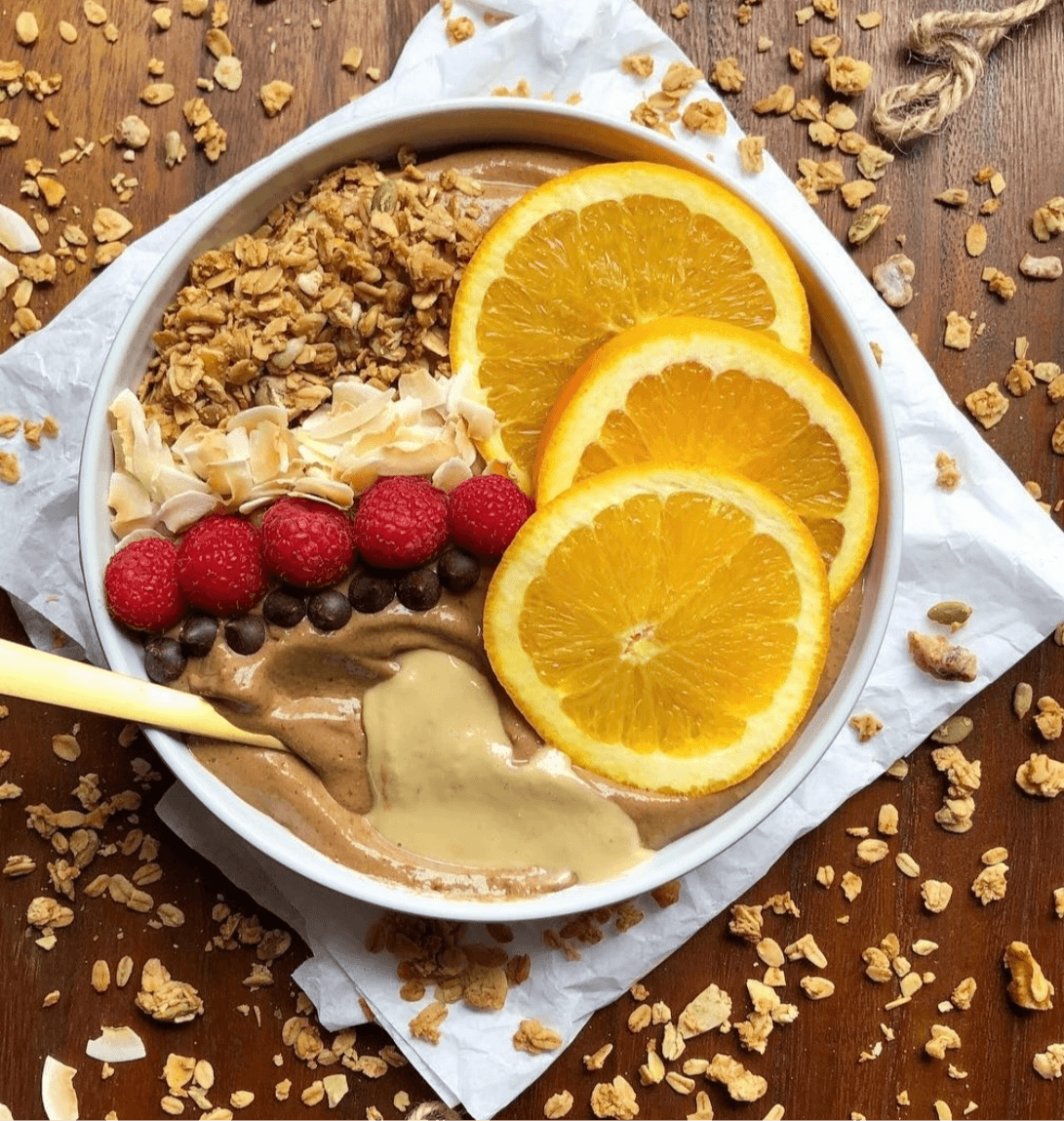 Bowl of dessert with orange slices, raspberries, granola, and chocolate topping on a wooden surface.
