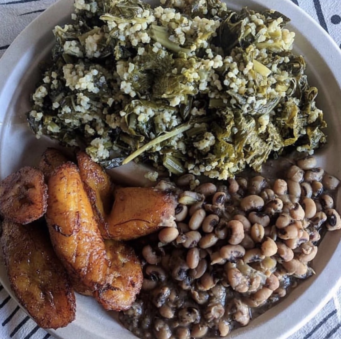Plate with cooked greens, black-eyed peas, fried plantains, and a side of rice and beans.