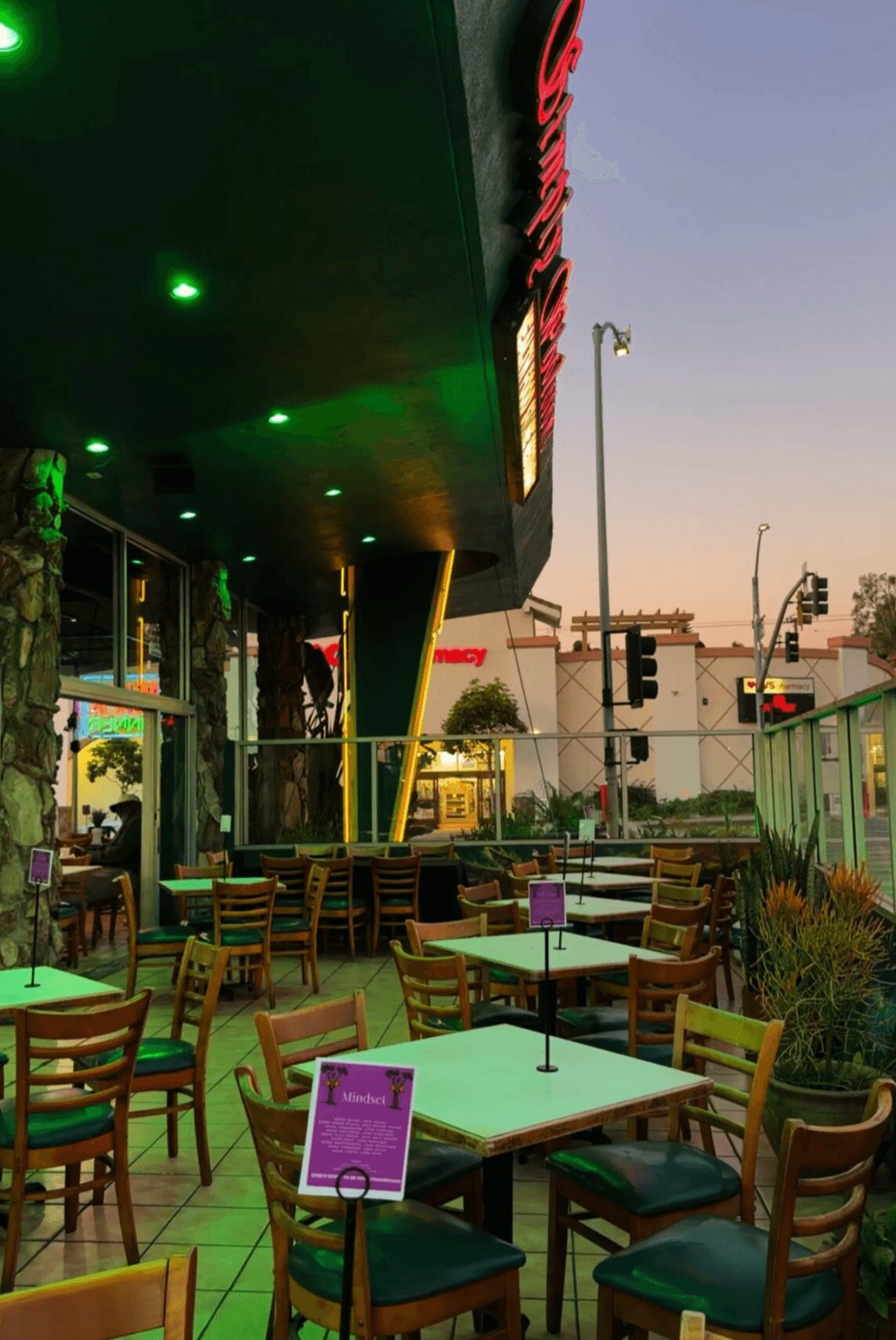 Outdoor seating area with tables and chairs outside a building with green lighting and a purple sign.