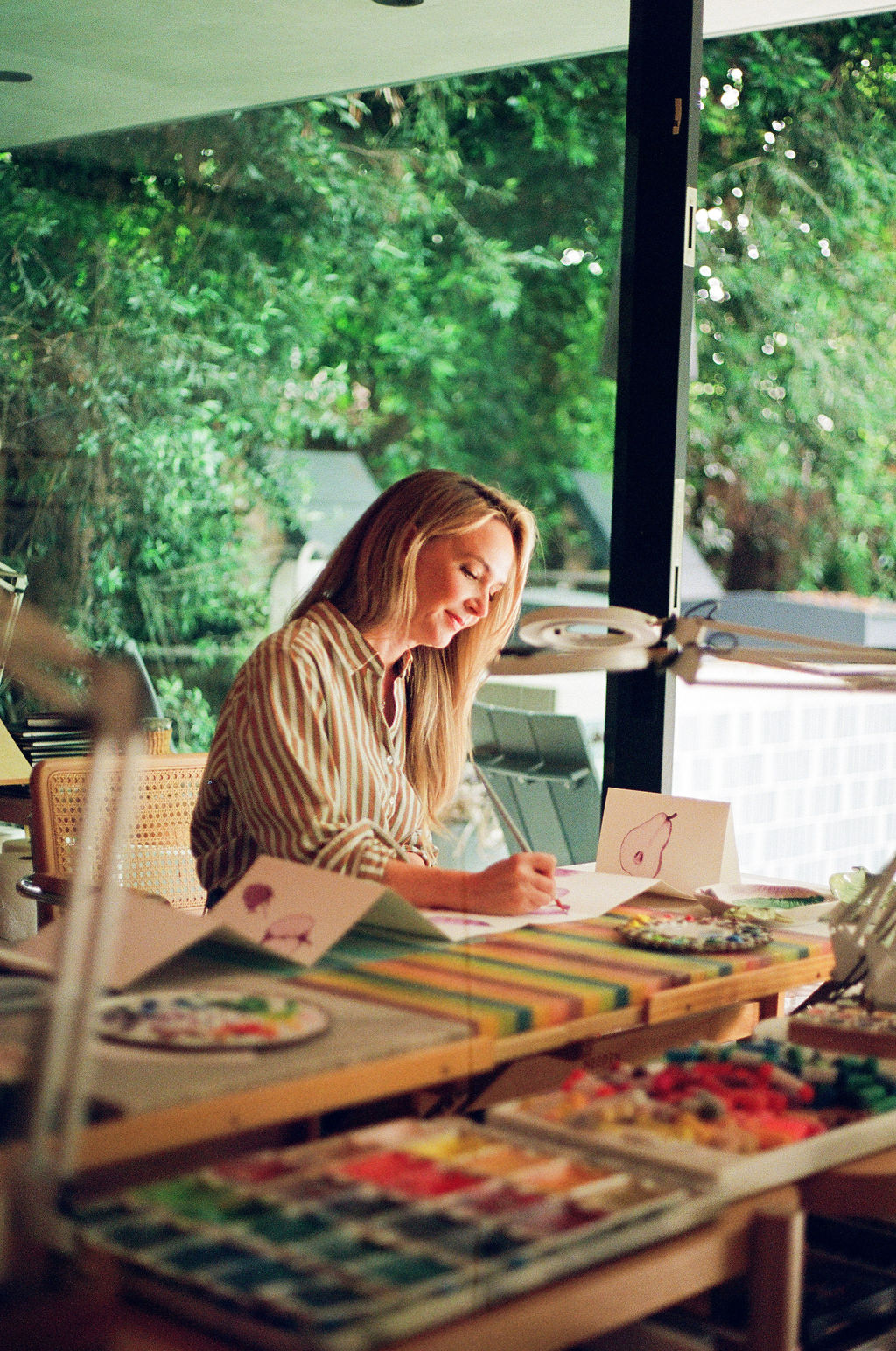 Woman sitting at a table with colorful items, writing on a paper, near large window with green trees outside.