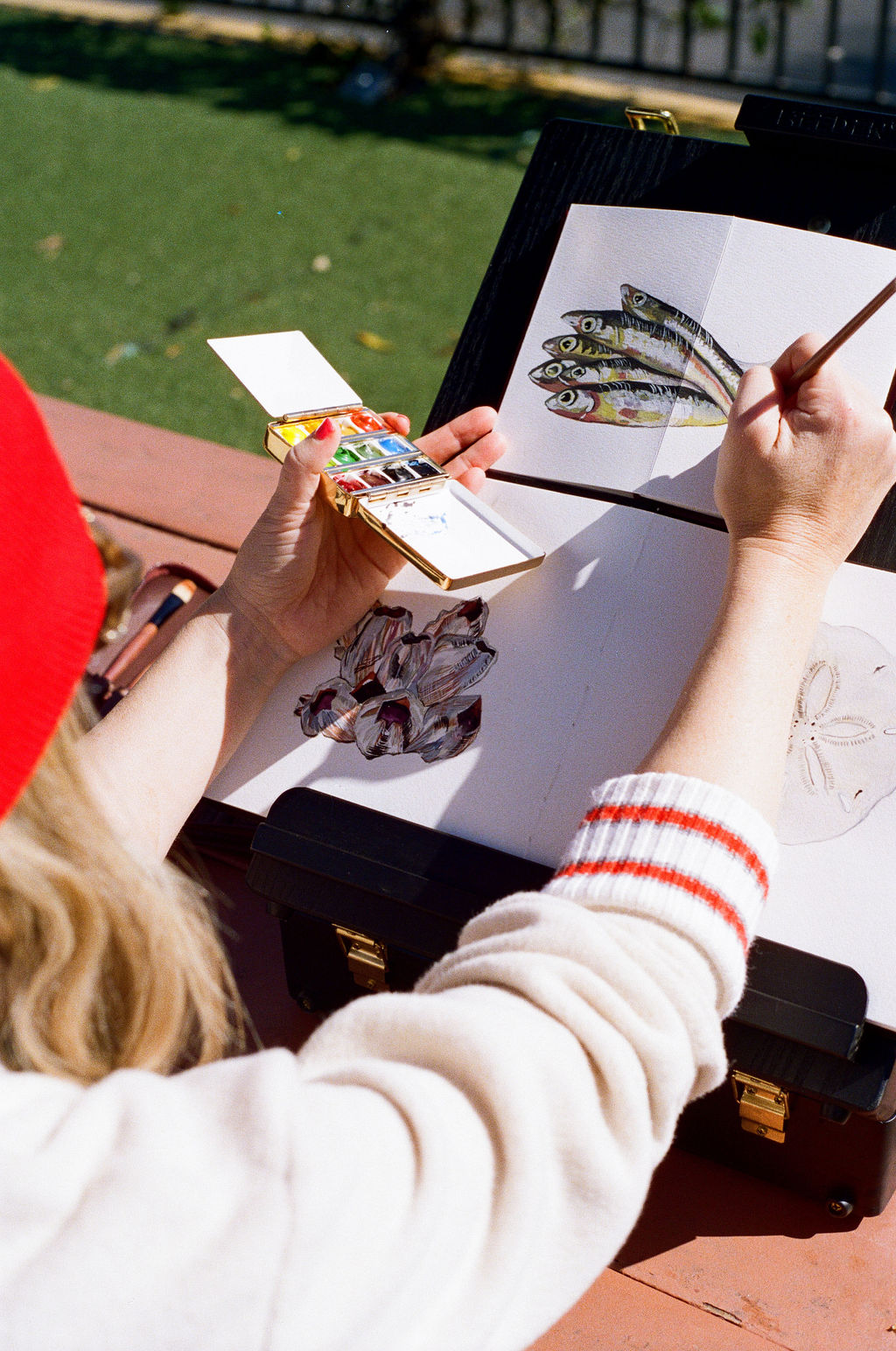 Child painting on paper with a paintbrush, outdoors on a red surface, with a palette in hand.