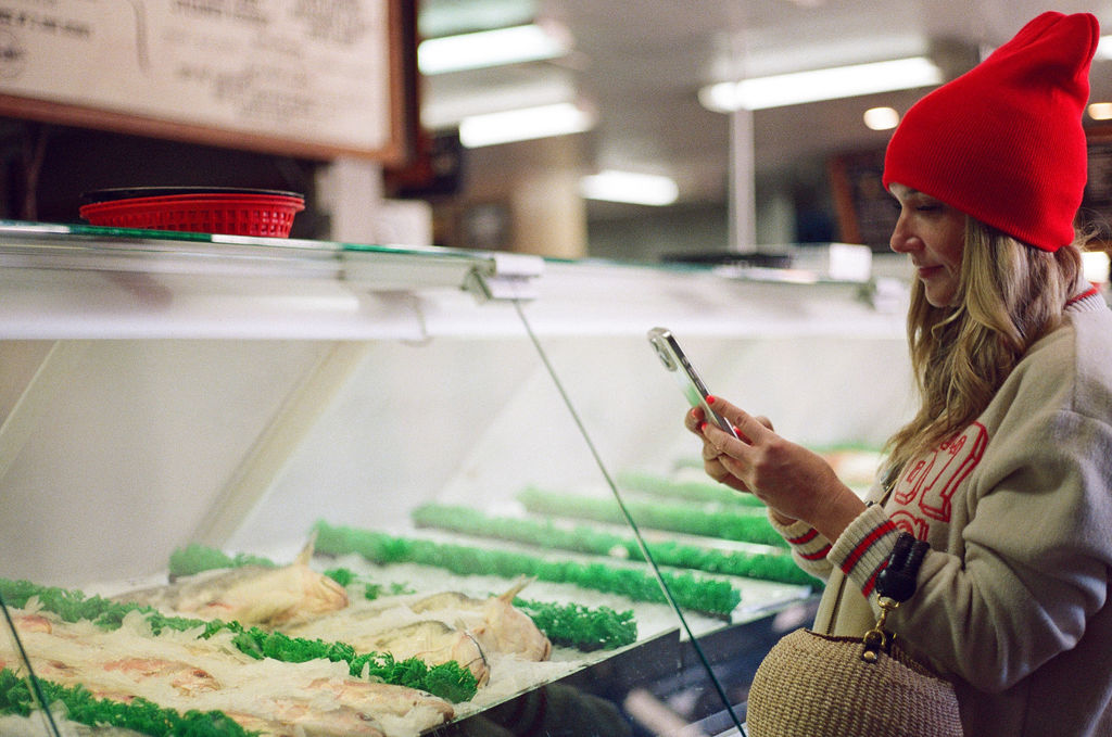Person wearing a red hat shopping for ice cream at a store, looking at their phone.