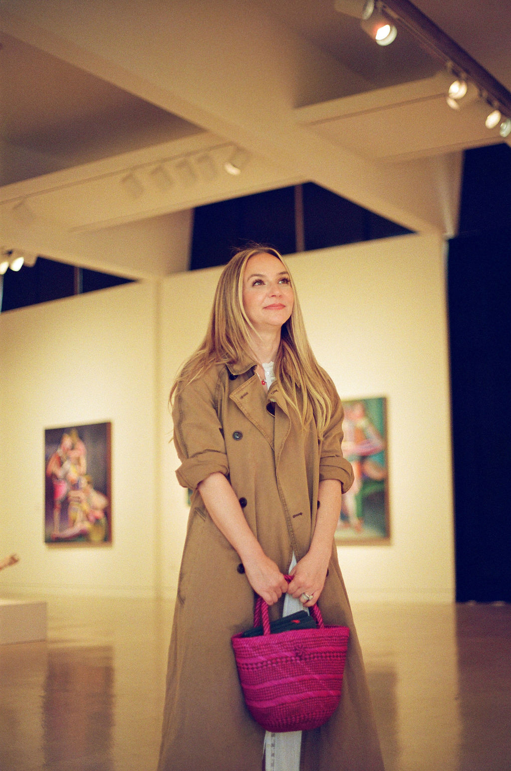Young woman with long blonde hair in an art gallery, holding a pink bag, looking upward.