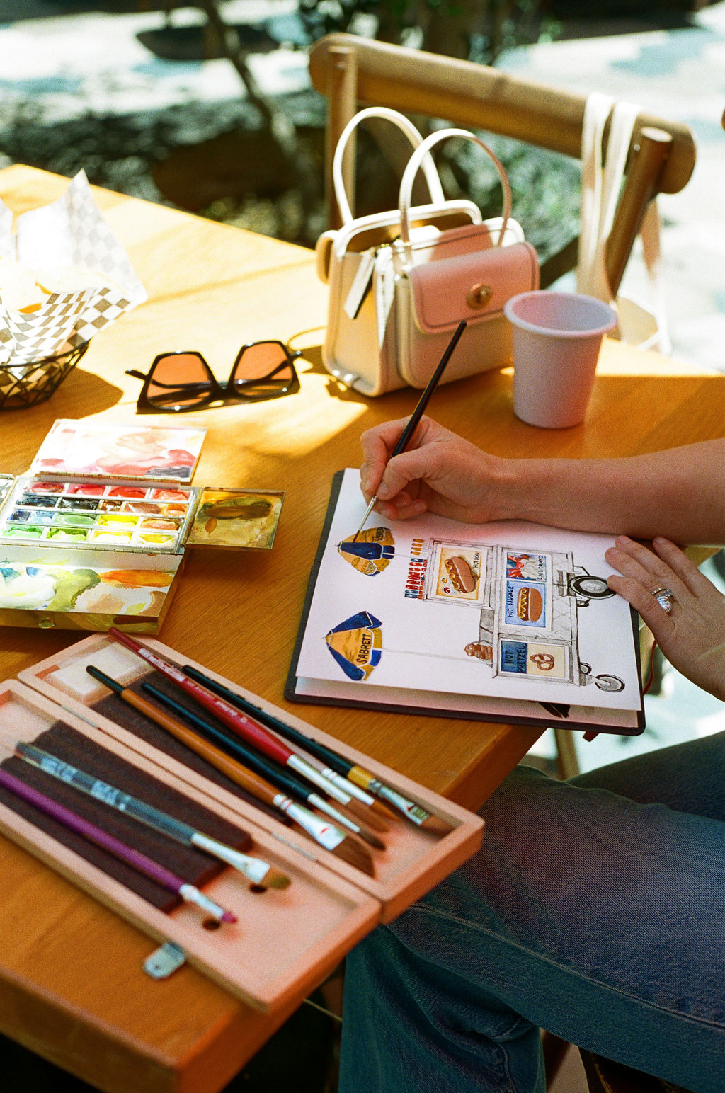 Person drawing on a sketchbook with colored charts, surrounded by art supplies, handbags, sunglasses, and a cup on a wooden table.