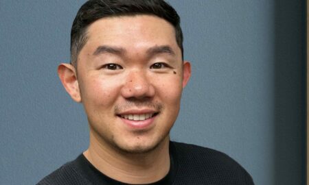 Young man with short black hair smiling, wearing a black shirt, standing against a blue wall.