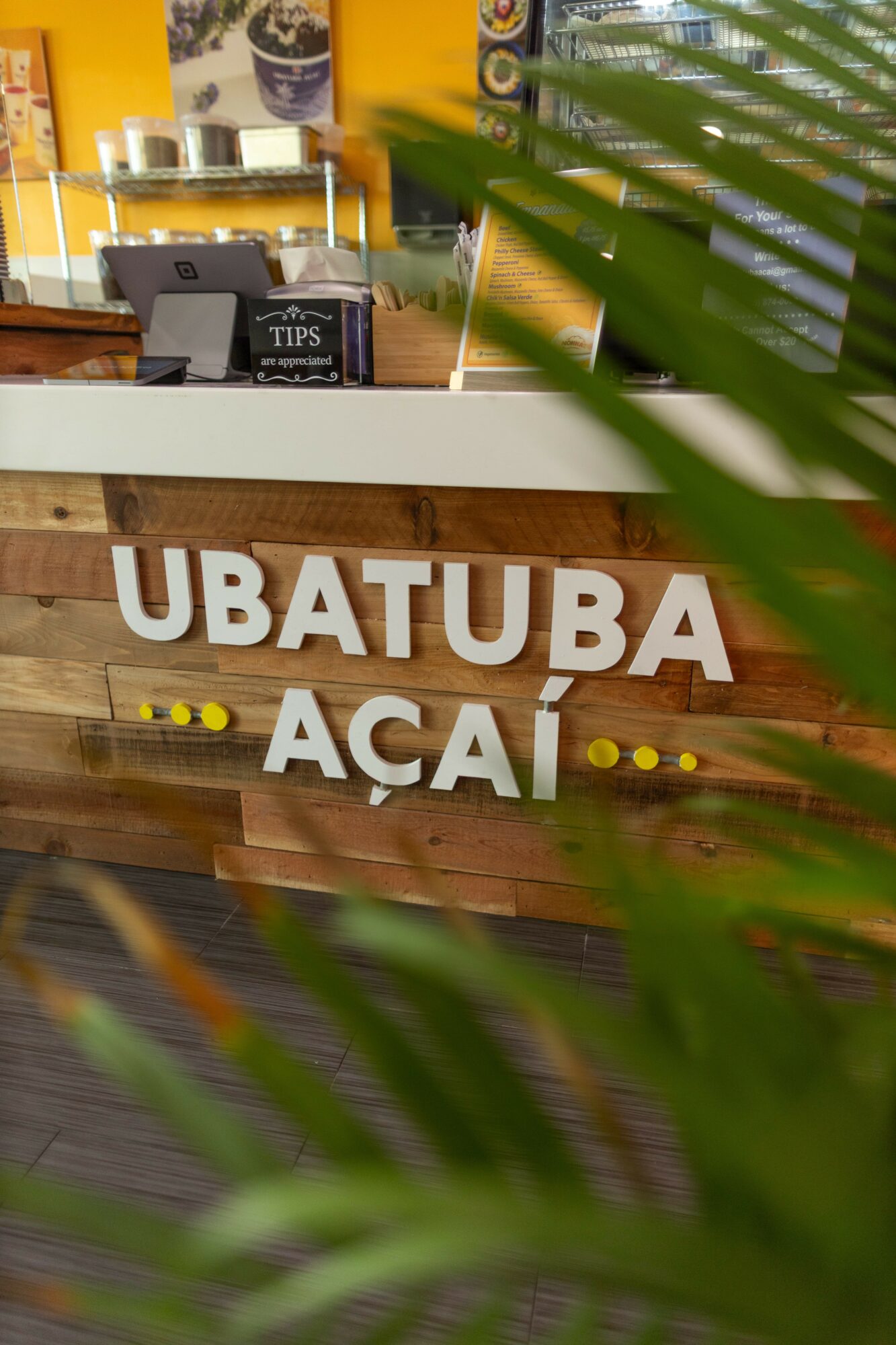 Sign with the words Ubatuba Açaí in front of a wooden counter, with a yellow wall and menu in the background.