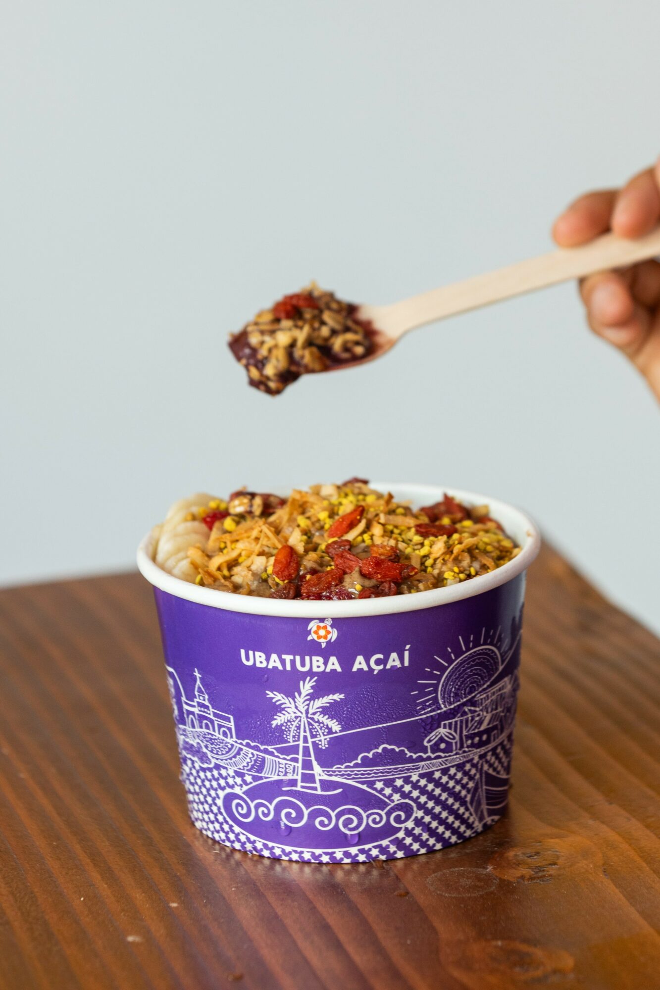 Hand holding a spoon with food above a purple container on a wooden surface.