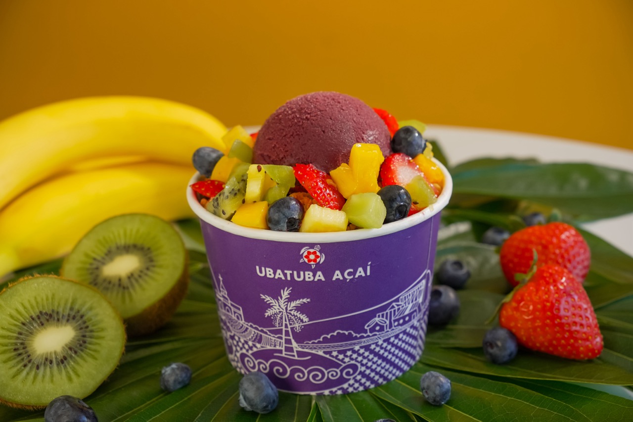 Cup of acai with mixed fruit, surrounded by bananas, kiwi, strawberries, and blueberries on green leaves.