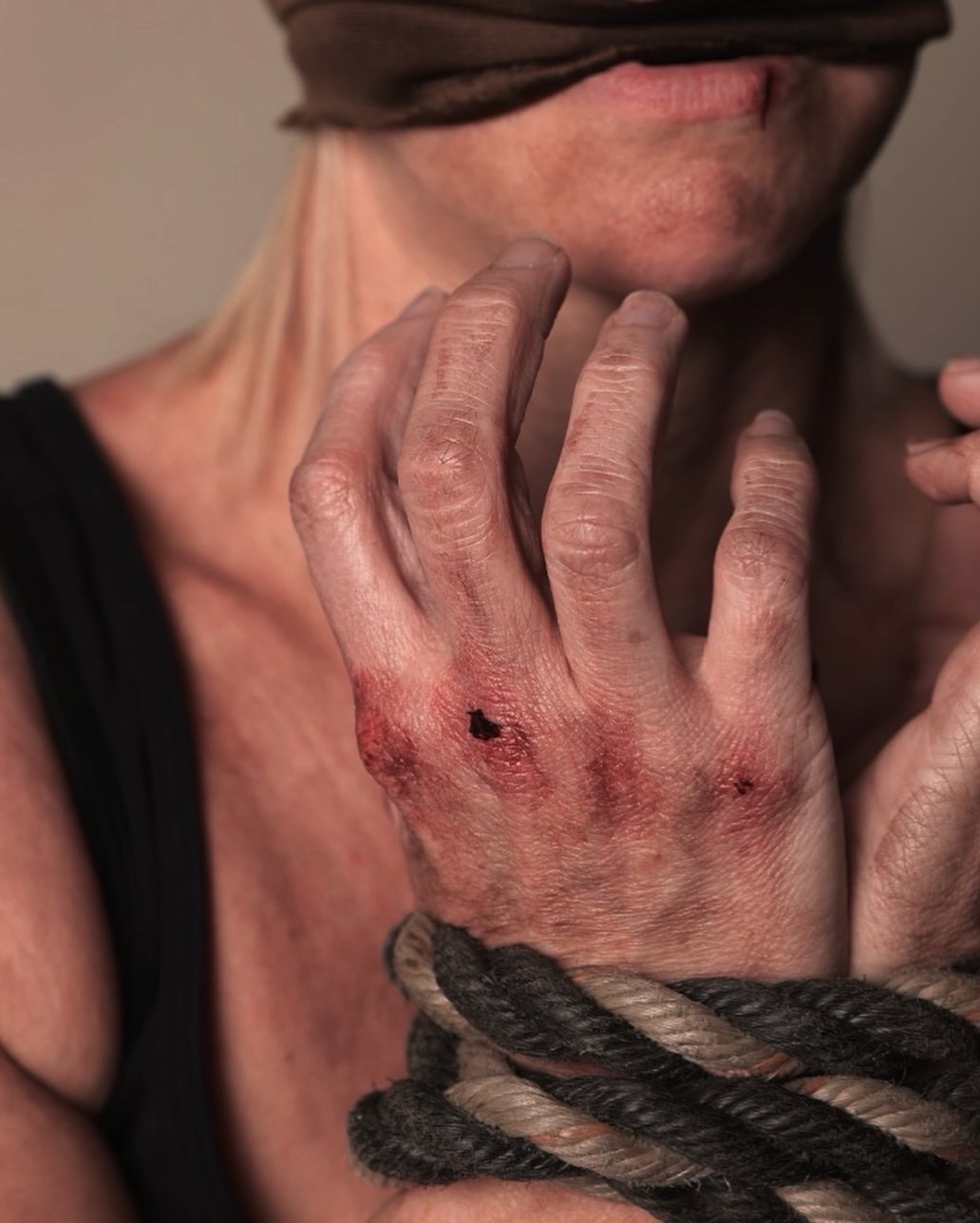 Close-up of a person's hand with visible wounds and a rope wrapped around the wrist, partially covering the face.