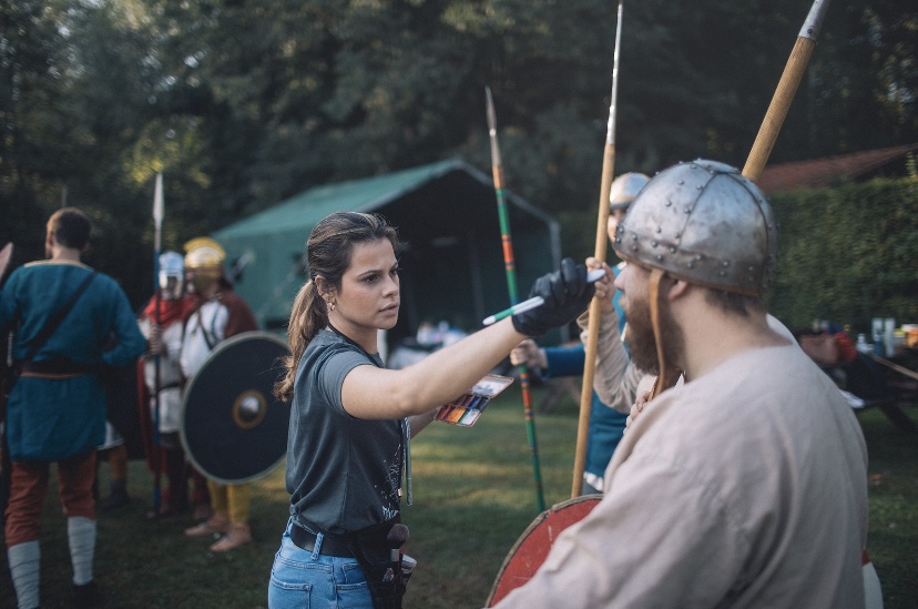 Woman in black shirt and jeans holding a sword, engaging with a man in medieval armor and helmet, outdoors.