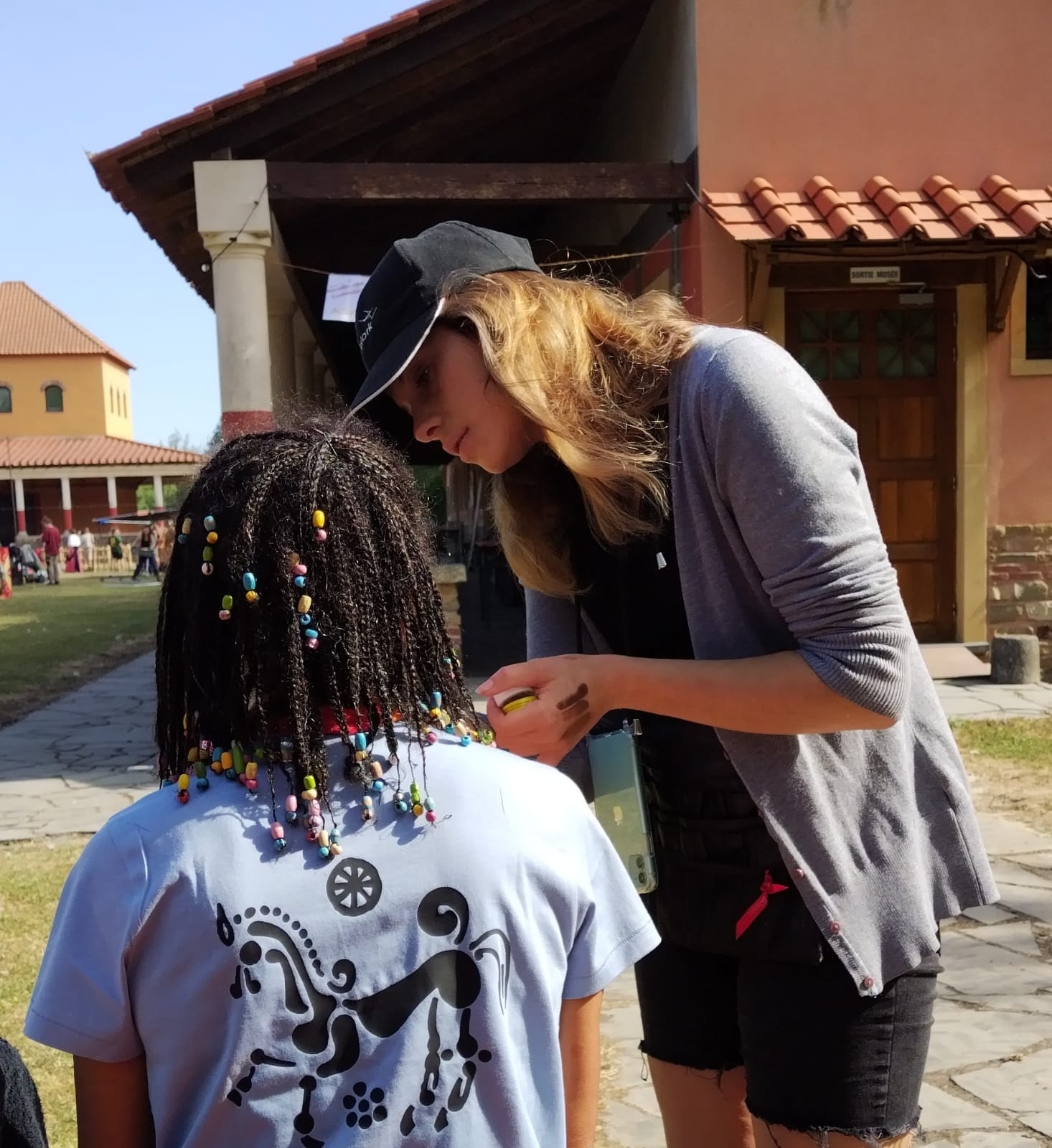 Two women engaged in conversation outdoors, one with curly hair and colorful beads in hair, the other with long blonde hair, wearing a cap.