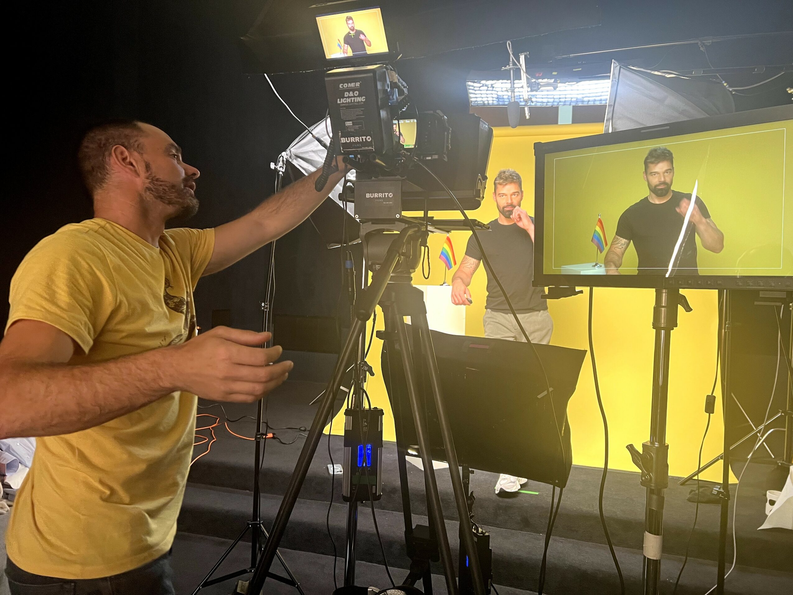 Two men in a studio with camera and monitor, one holding a microphone, yellow background, recording or live streaming setup.
