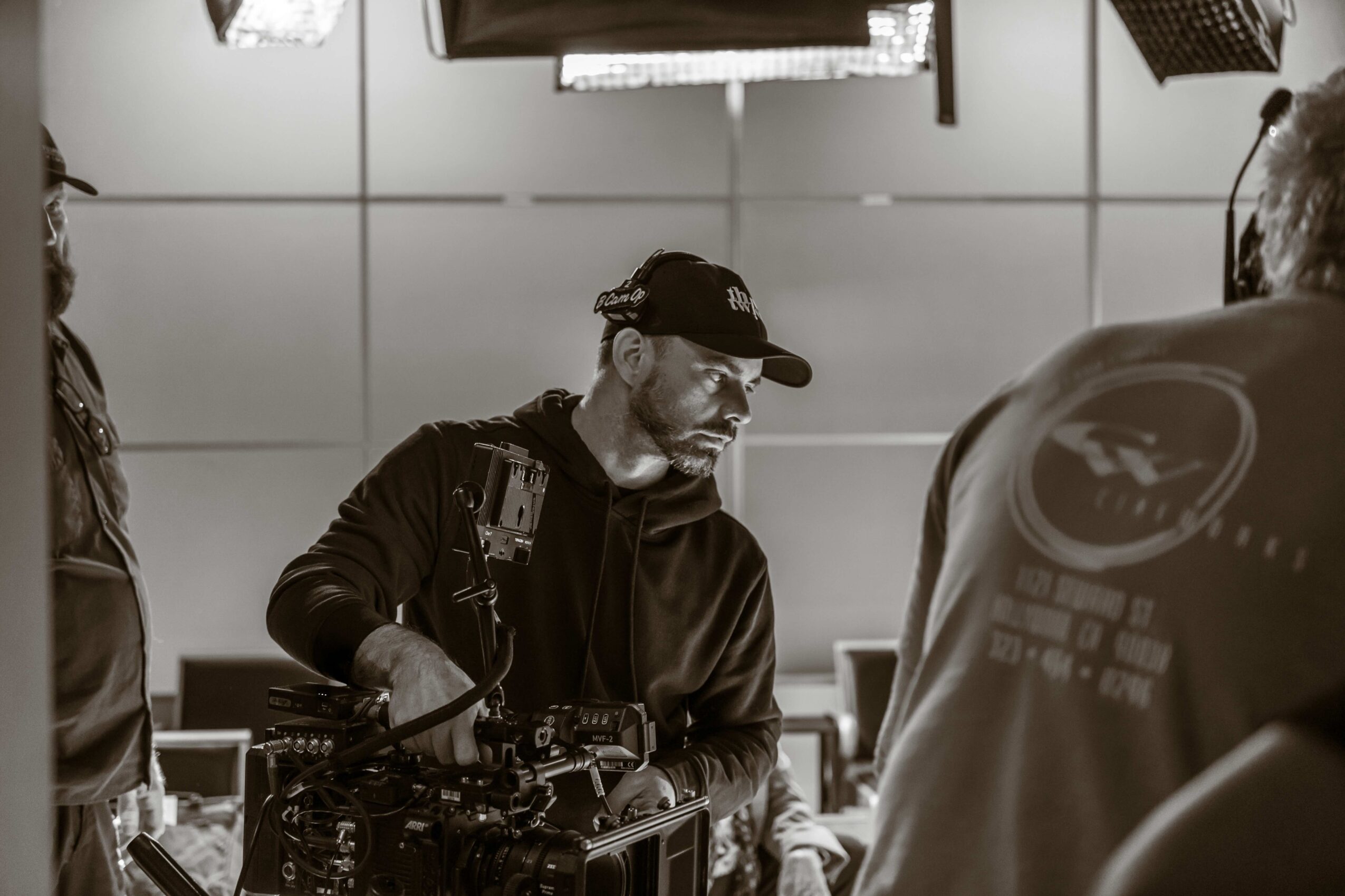 Man with a beard, wearing a cap and headphones, operating audio equipment in a room with a ceiling light panel.