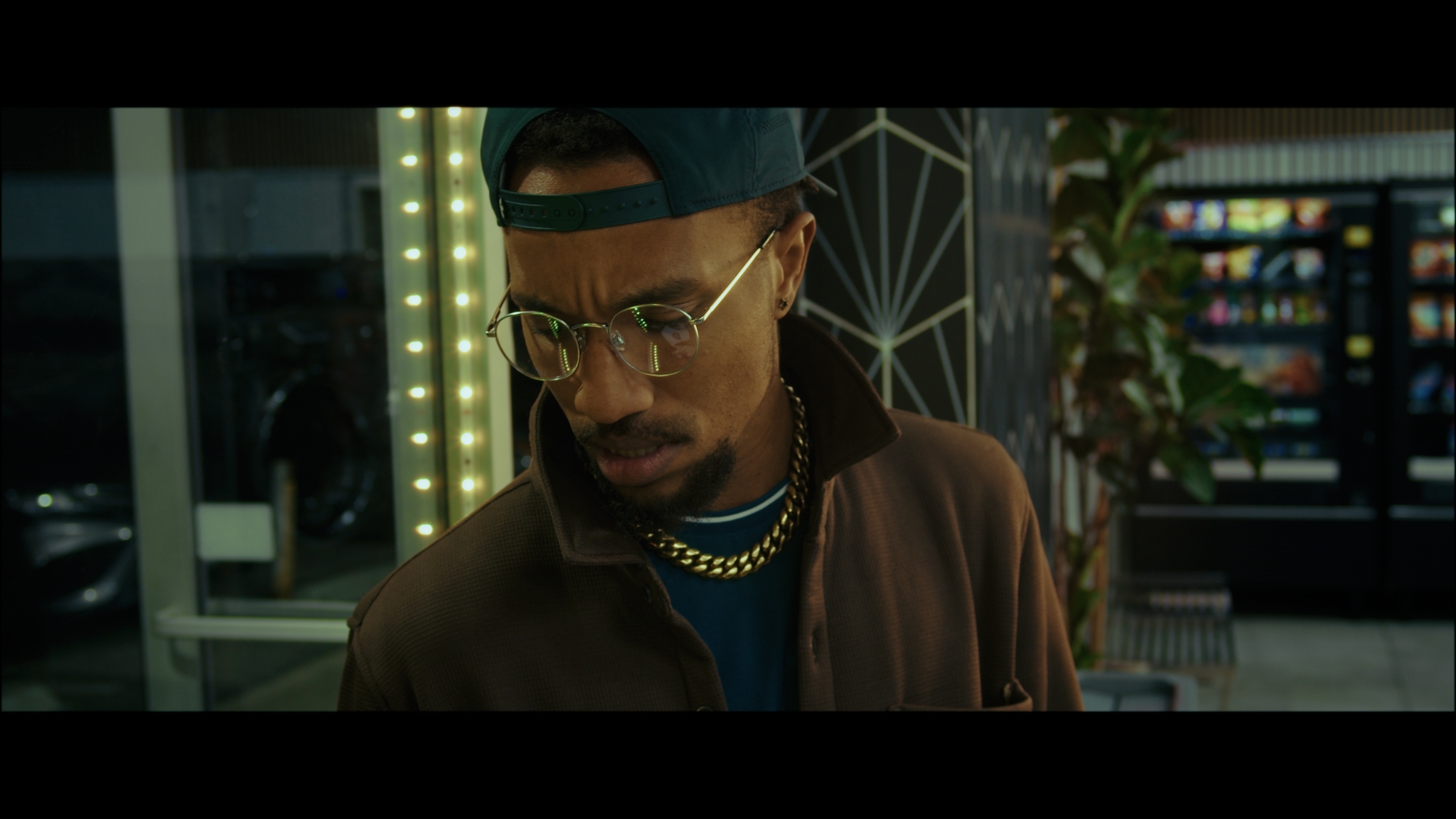 Man with glasses, cap, and gold chain looking down indoors near vending machines and plants.