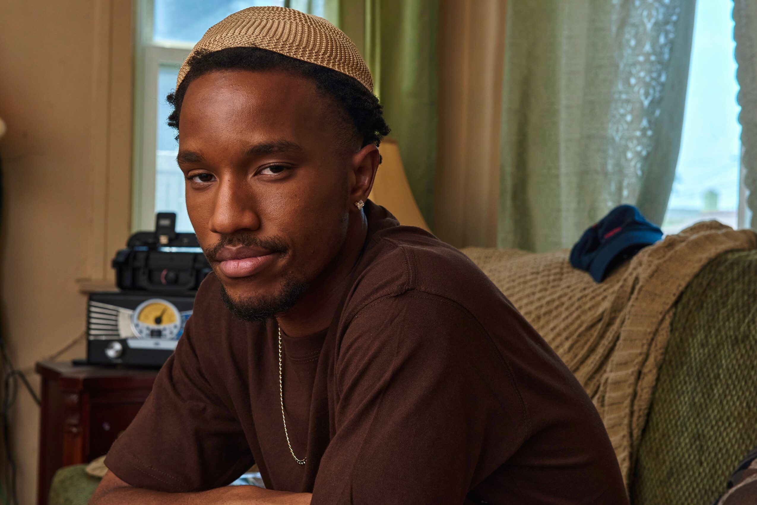 Young man with dark skin and braided hair, sitting on a couch in a room with green curtains and a window.