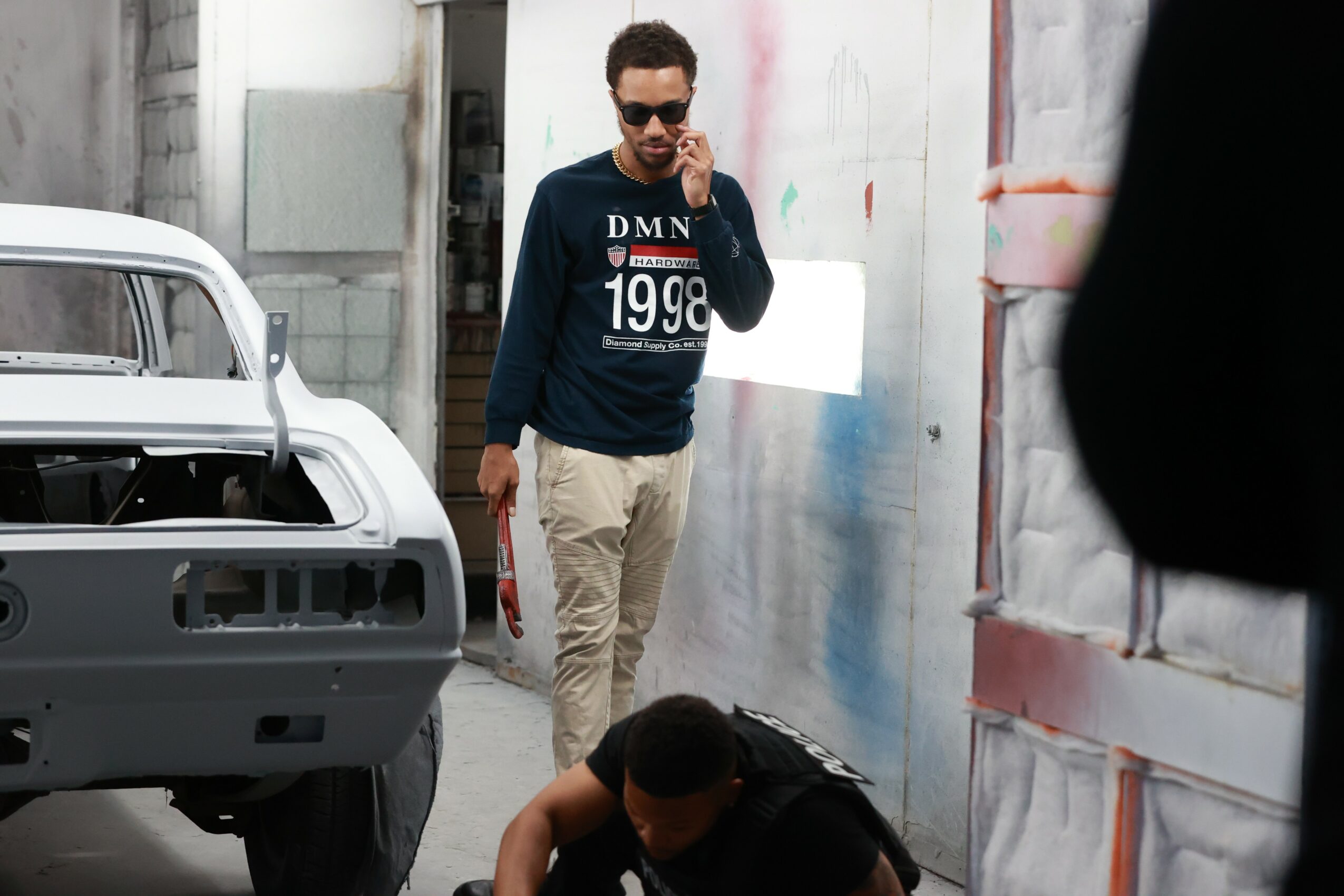 Man with sunglasses and a beard talking on a phone, standing near a white vehicle in an urban setting.