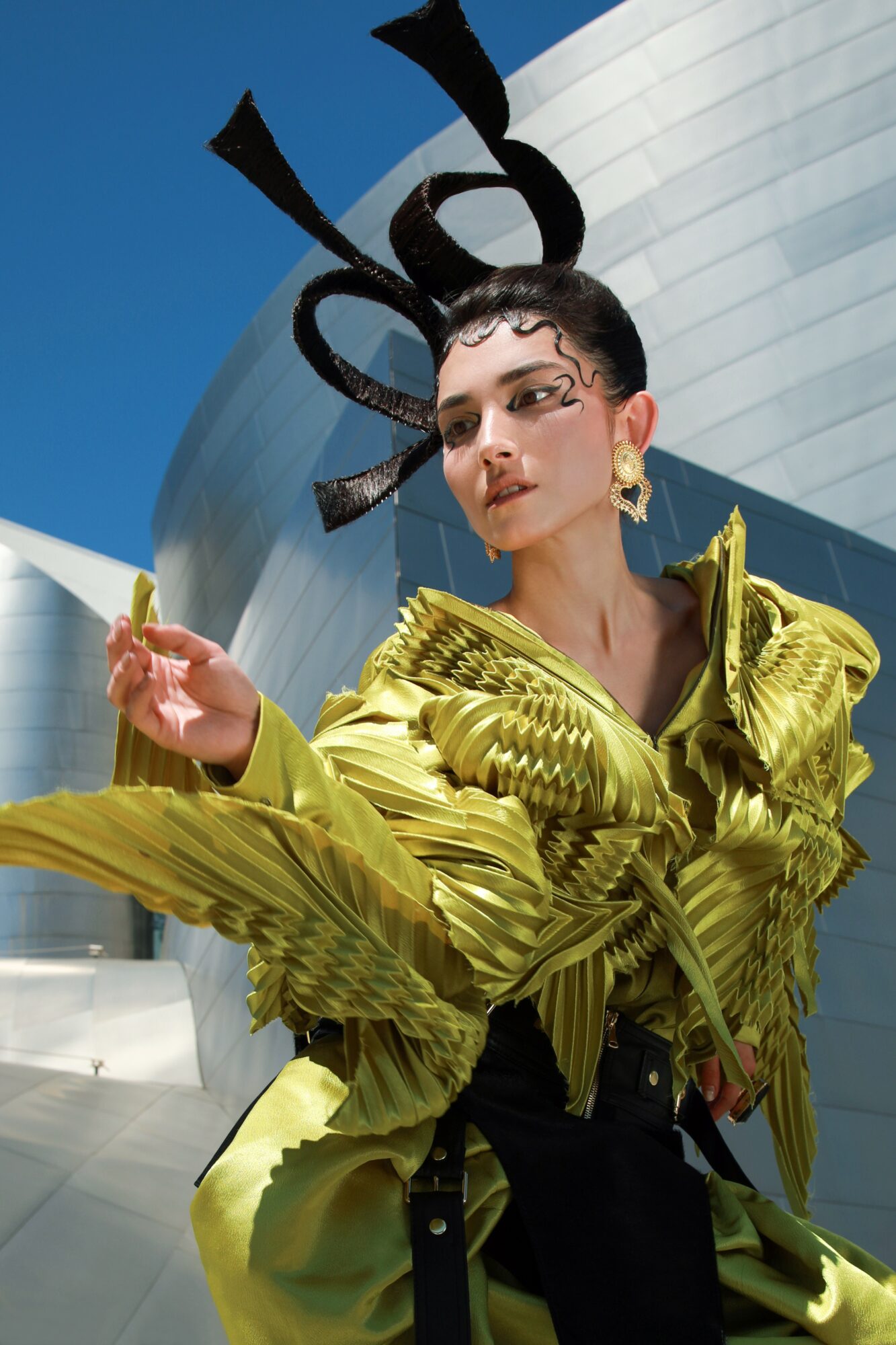 Person with elaborate headpiece and gold ruffled jacket outdoors, modern building in background.