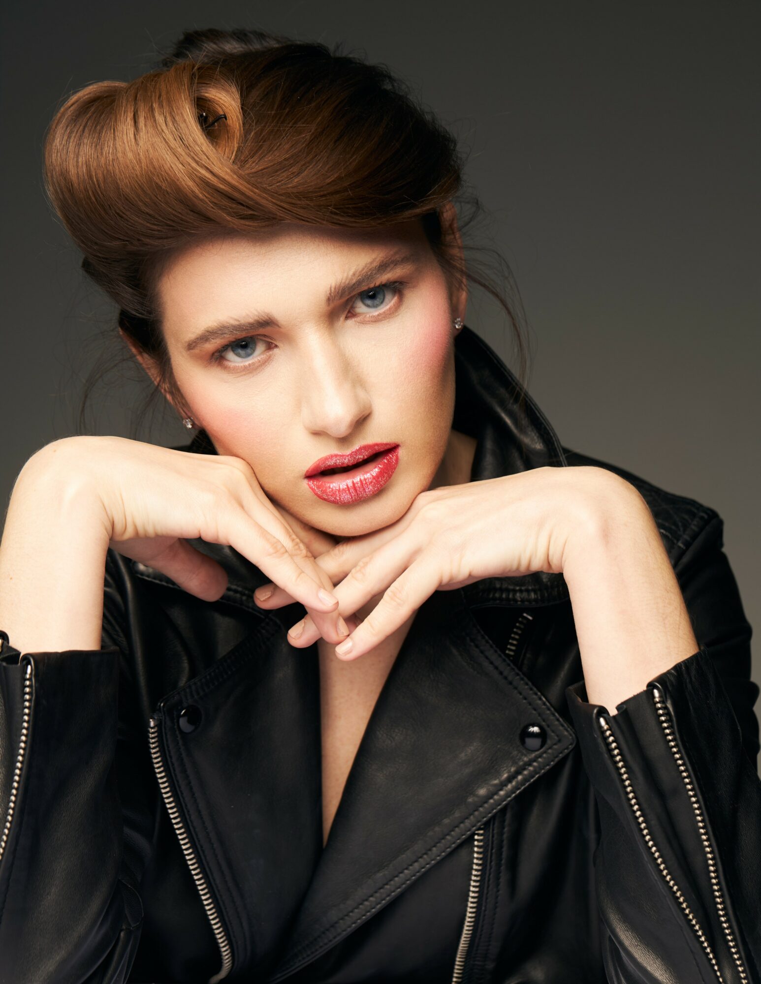 Woman with styled hair and makeup wearing a black leather jacket, posing with hands near face.