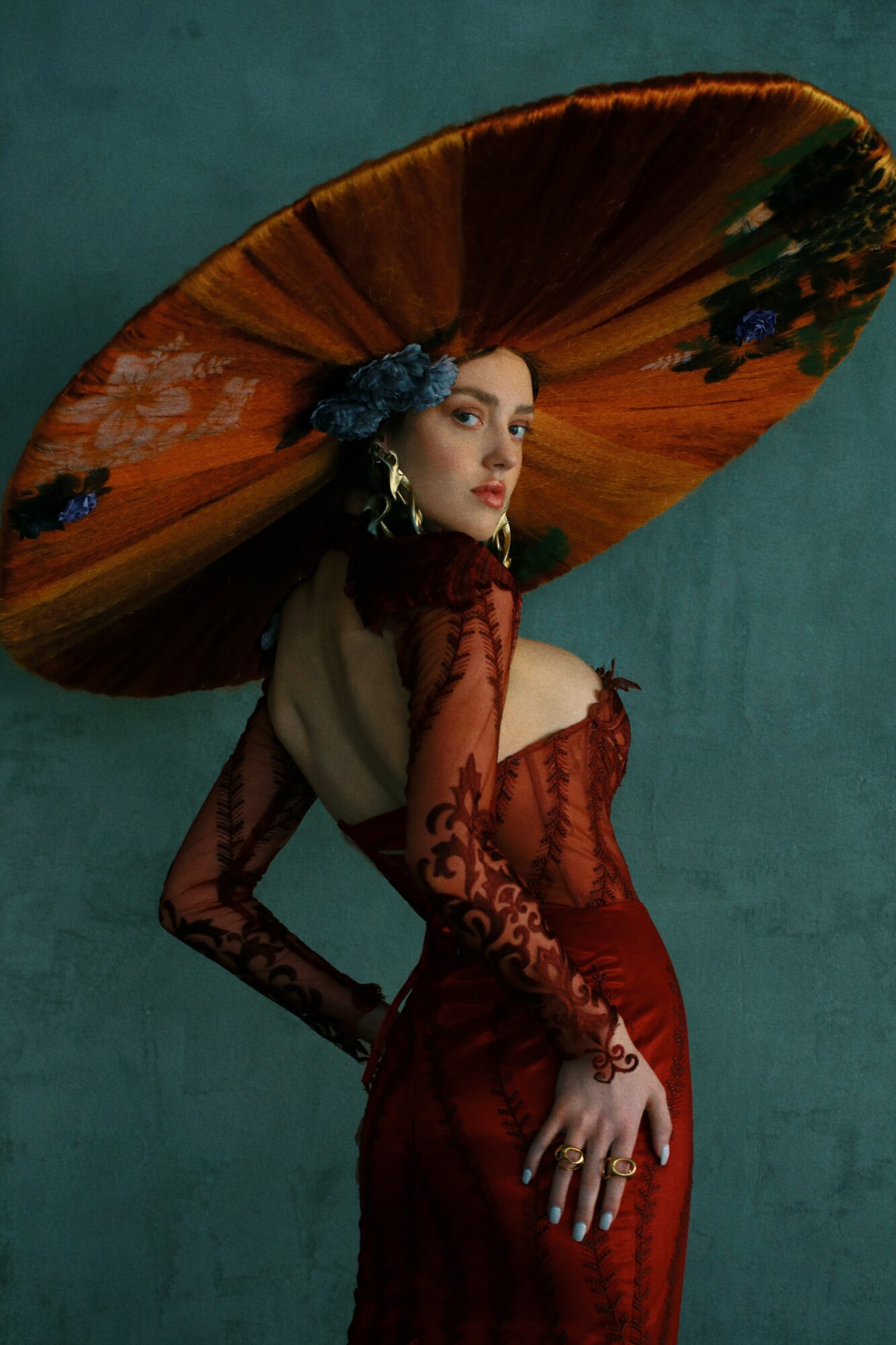 Woman in red dress with large, colorful umbrella on head, standing against a teal background.