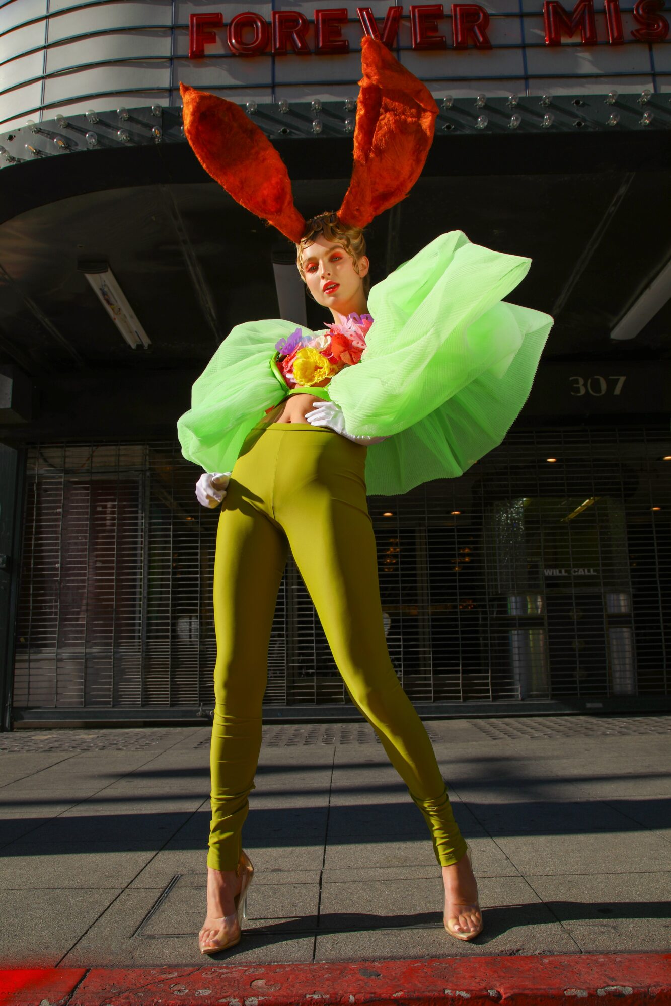 Person in colorful costume with large green sleeves and red bunny ears standing outdoors in front of a building.