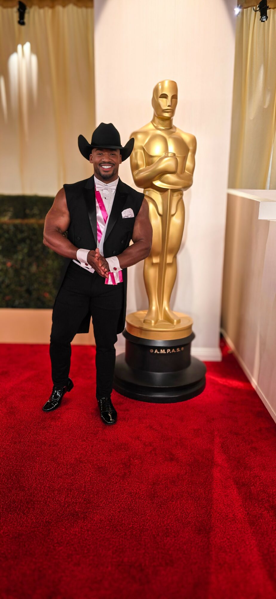 Man in black suit, pink tie, and black hat standing next to a gold statue on a black pedestal, red carpet background.