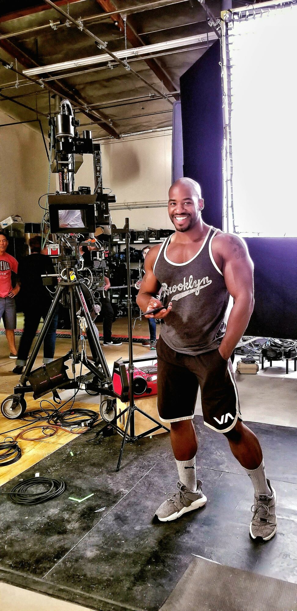 Man smiling in a gym, holding a phone, surrounded by camera equipment and gym gear.
