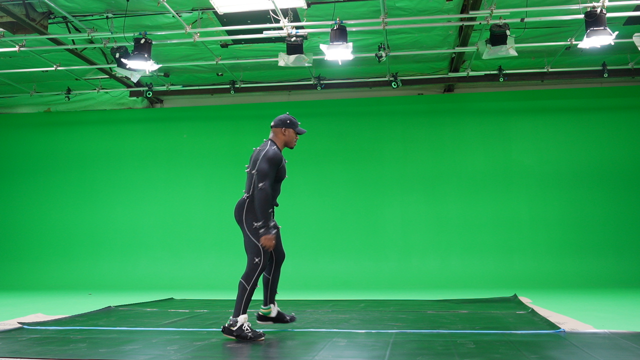 Person standing on a green screen studio floor, facing left, wearing athletic clothing and a cap.