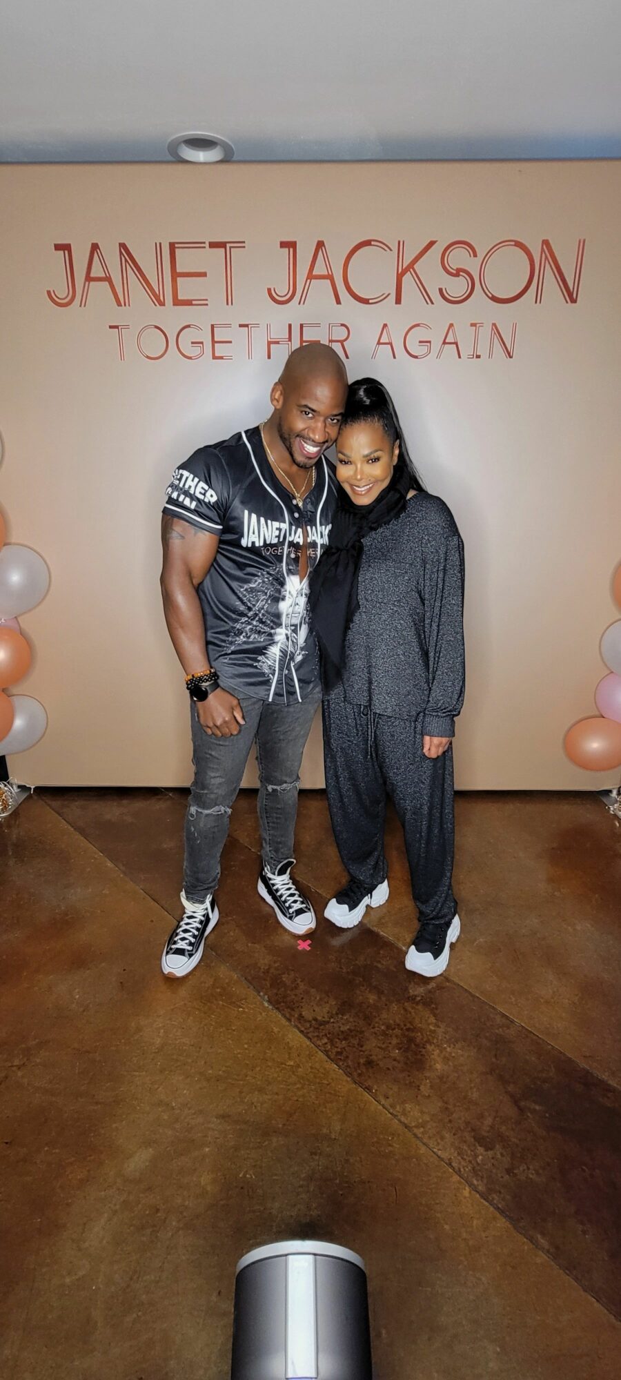 Two people standing together in front of a Janet Jackson backdrop with balloons on each side.