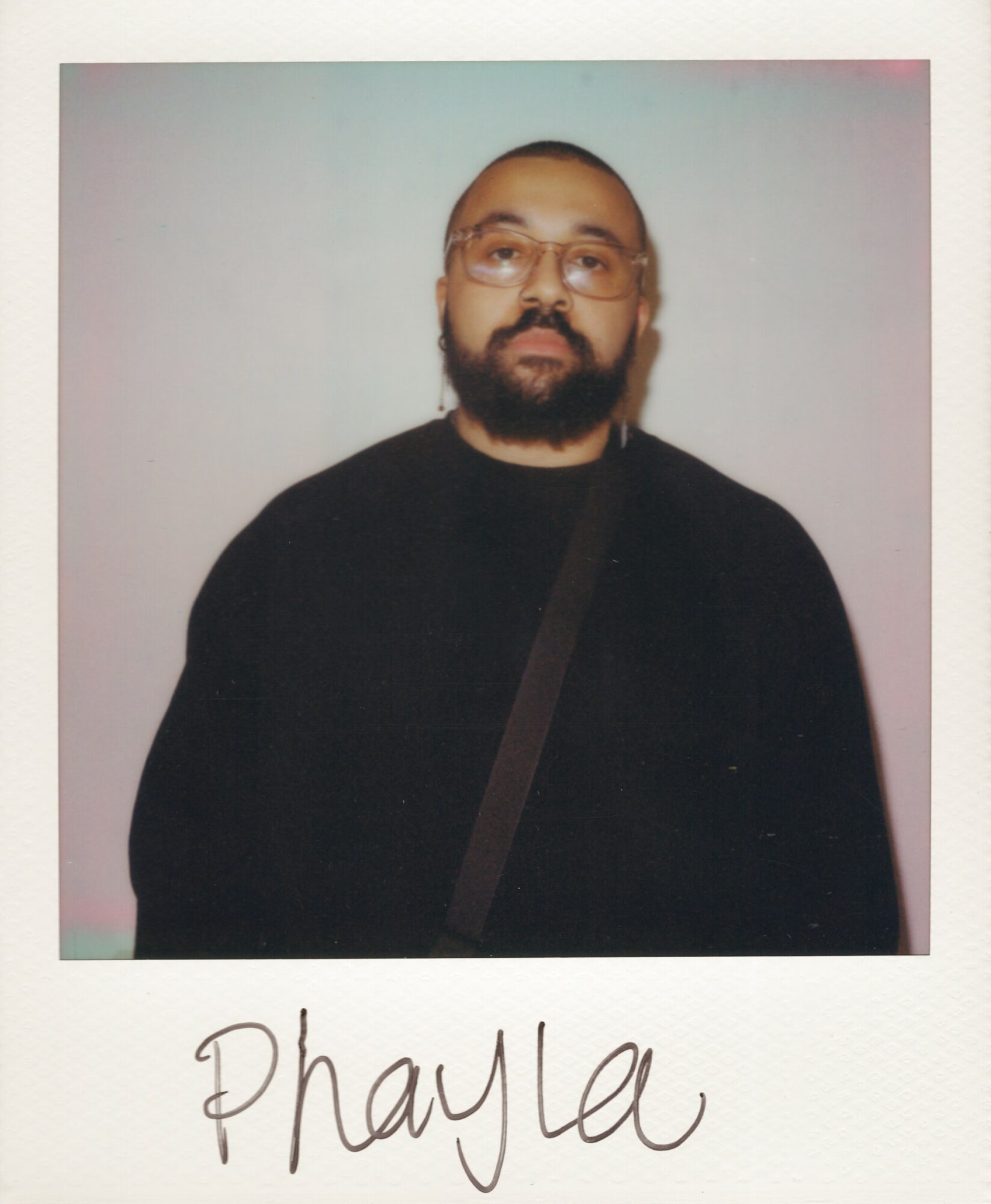 Person with glasses and beard wearing a black top, carrying a strap, against a plain background.