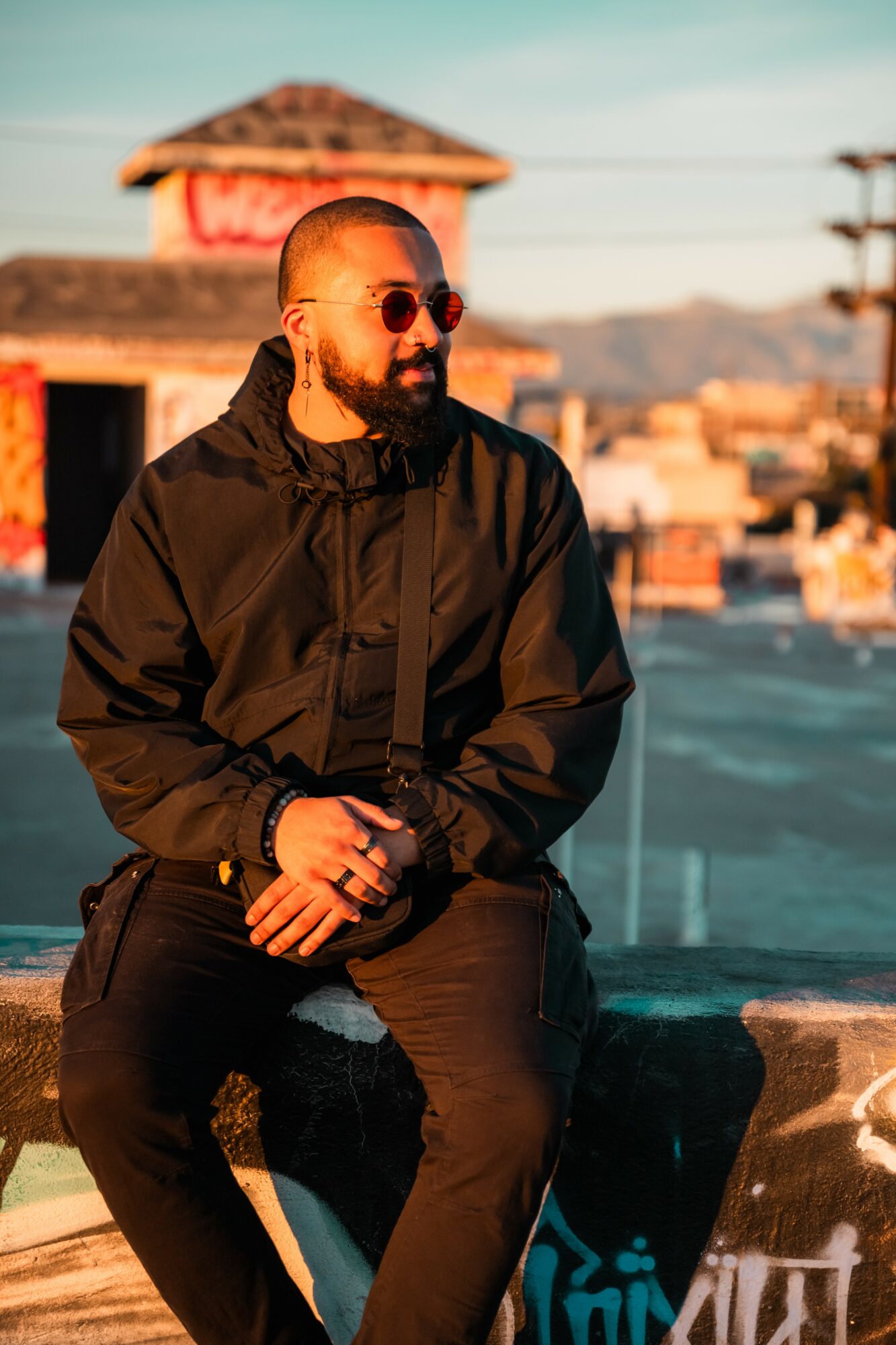 Man with sunglasses and beard sitting outdoors on a ledge, cityscape and mountains in background.