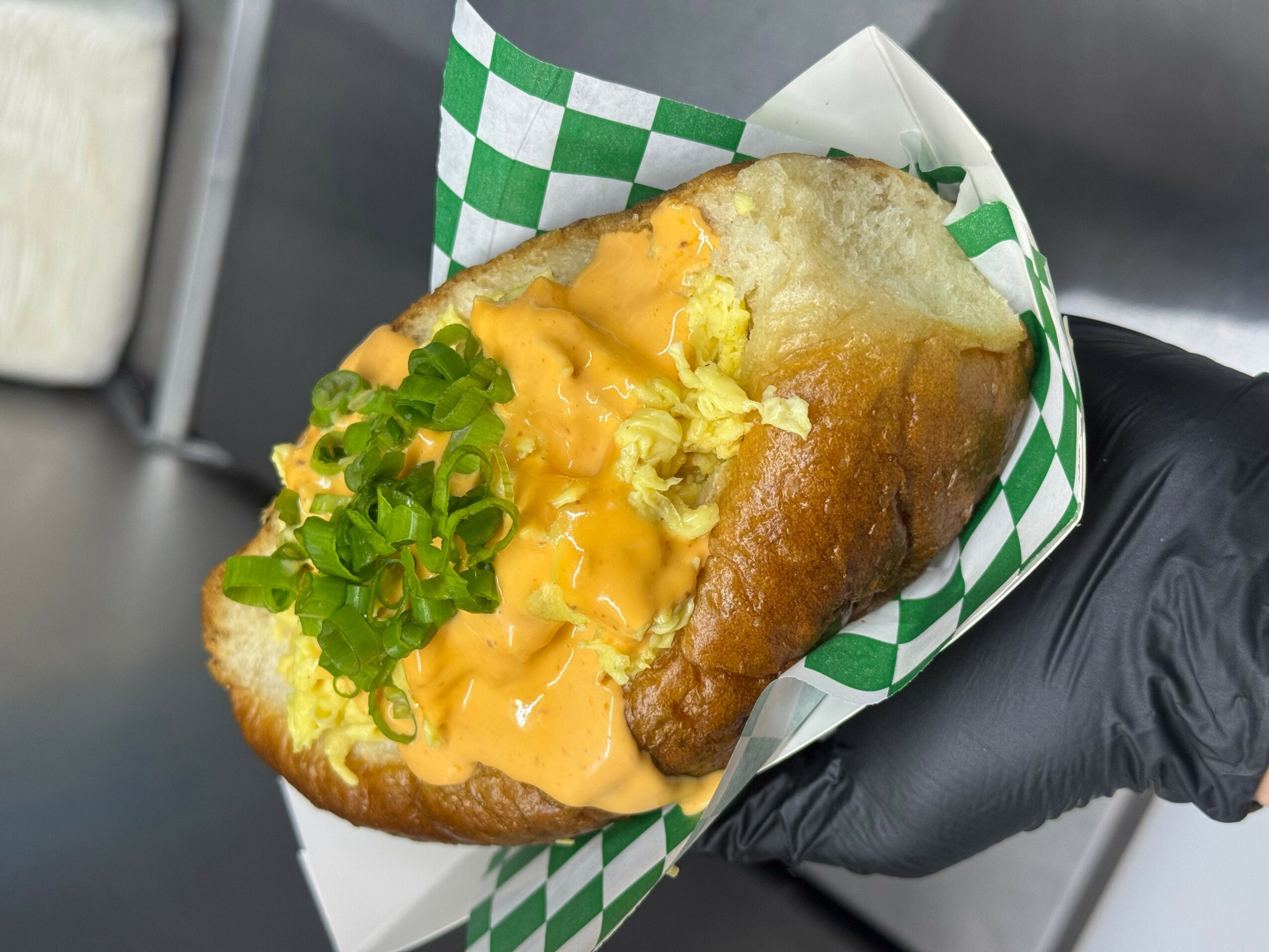 Hot dog with mustard and chopped green onions in a paper wrapper, held by a gloved hand.