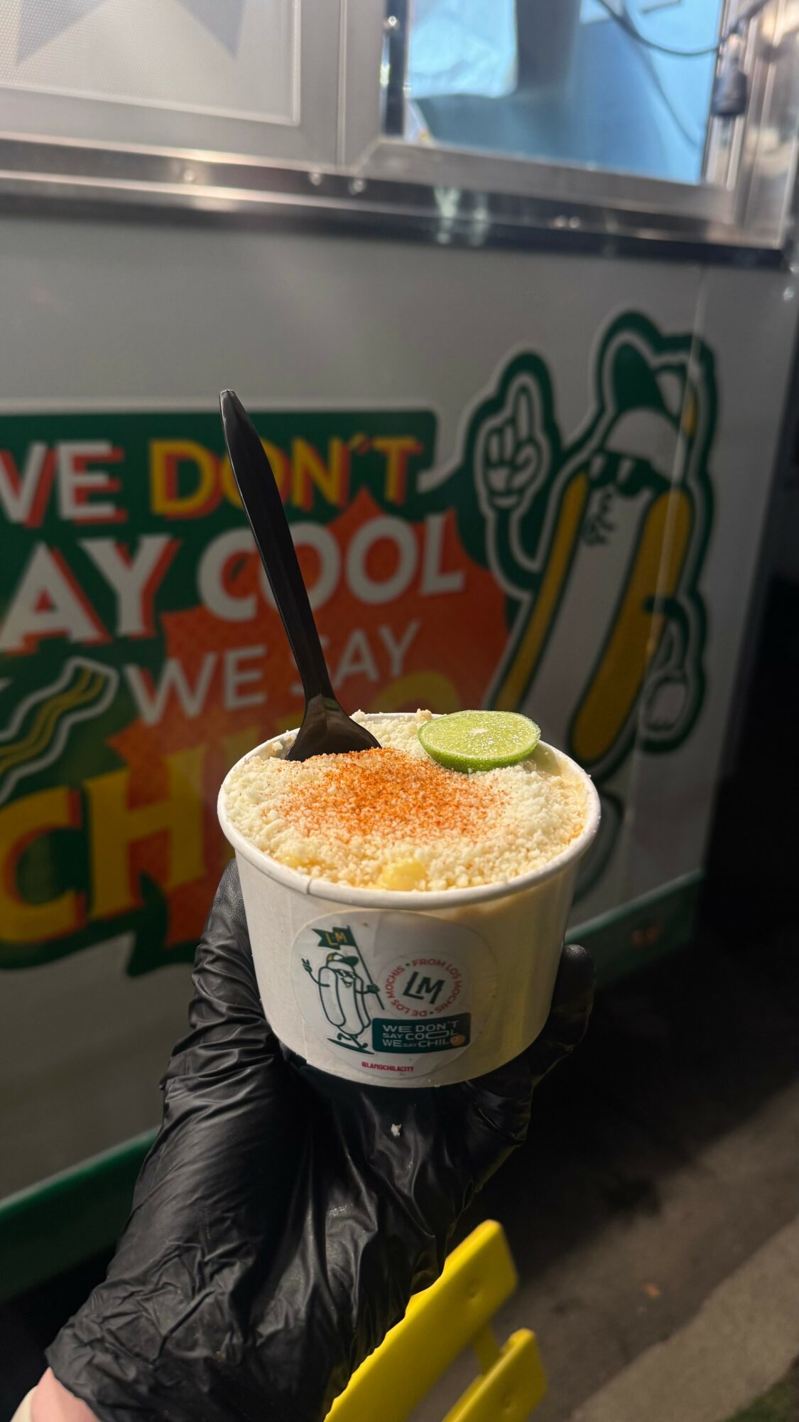 Cup of shaved ice with lime slice and syrup, held by a gloved hand in front of a colorful sign.