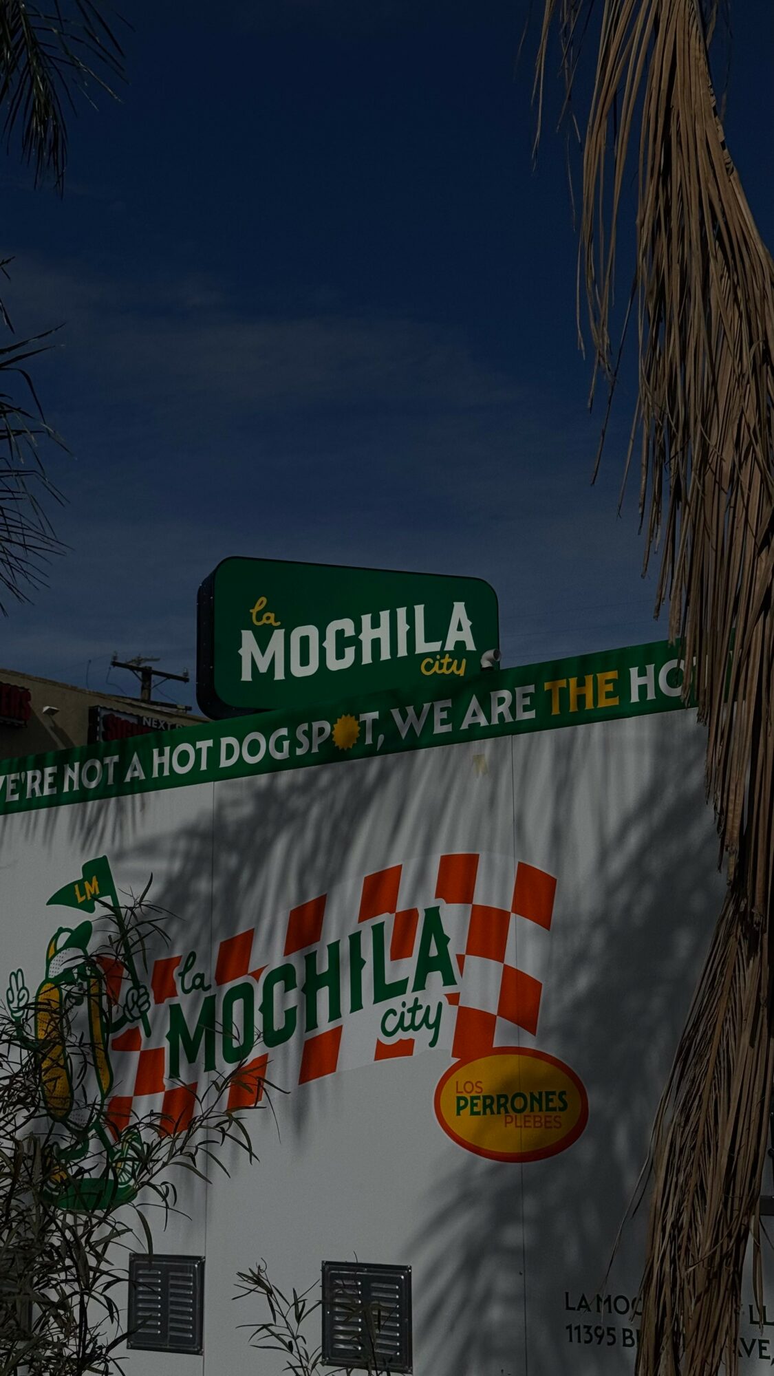 Signboard with the text 'MOCHILA' and a logo, partially obscured by tree branches, against a dark sky.