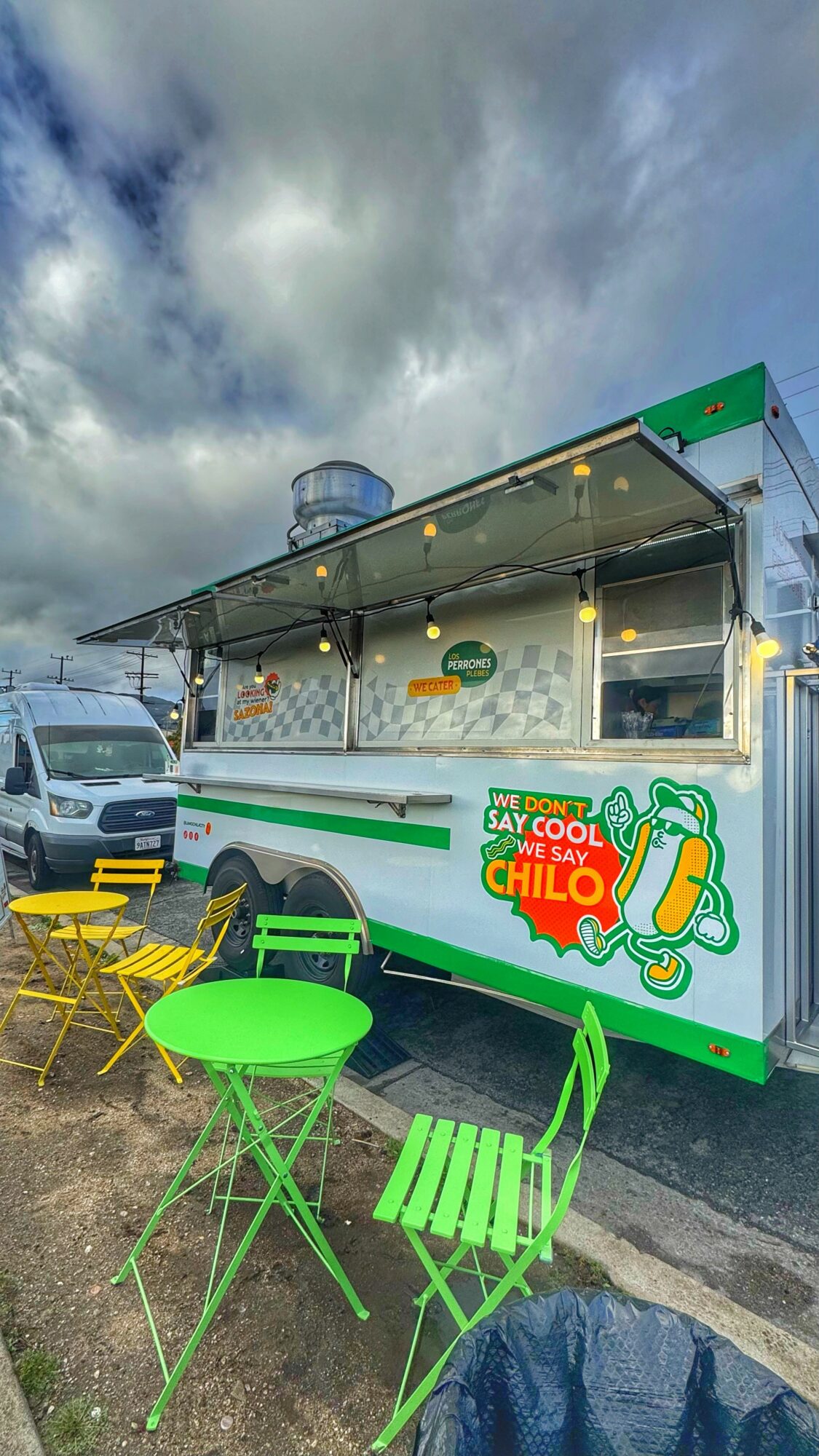 Food truck with yellow and green chairs outside, cloudy sky background, serving window open, colorful sign on side.