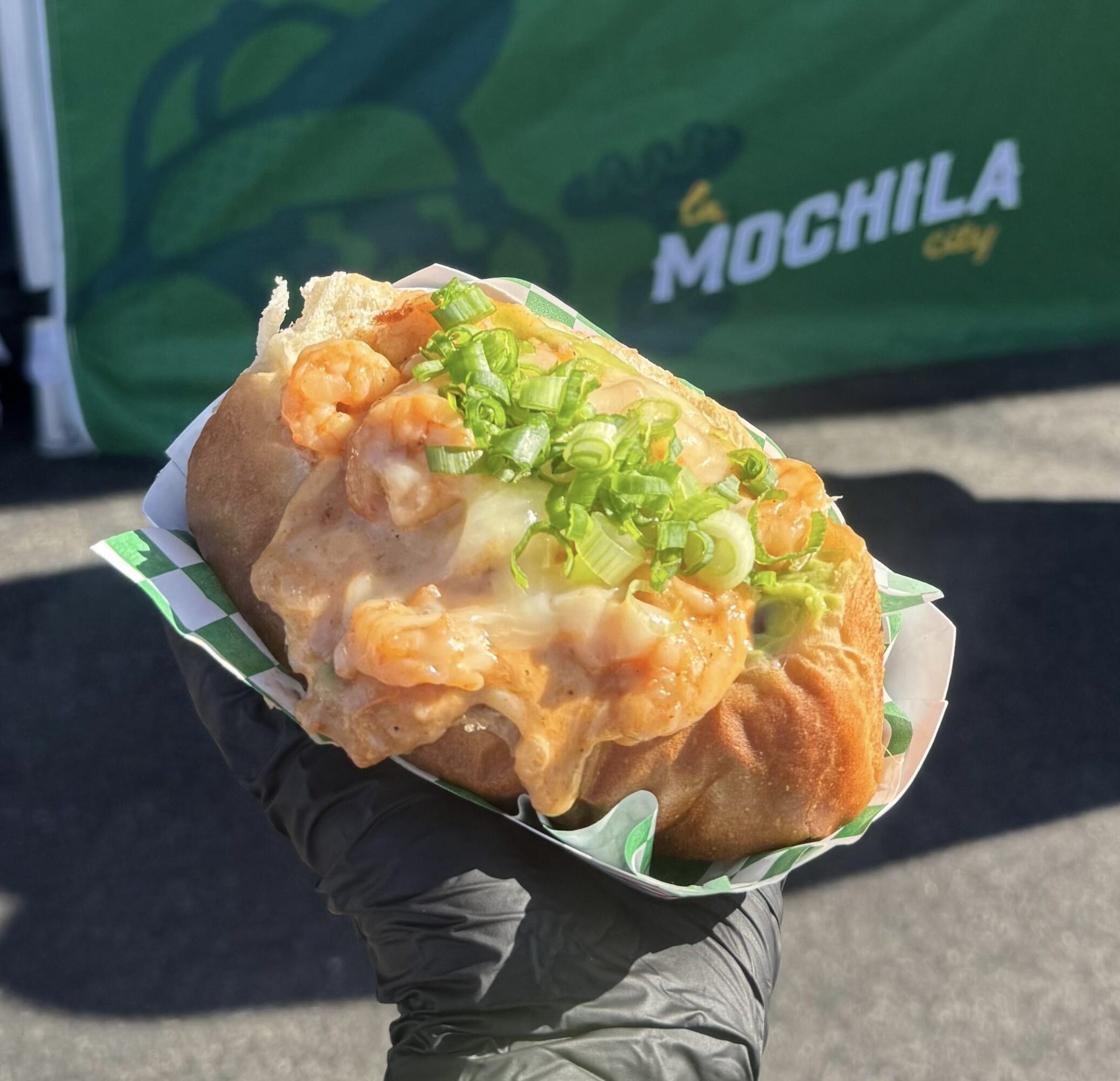 Hand holding a fried snack topped with green onions, in a paper tray, outdoors with a green background.