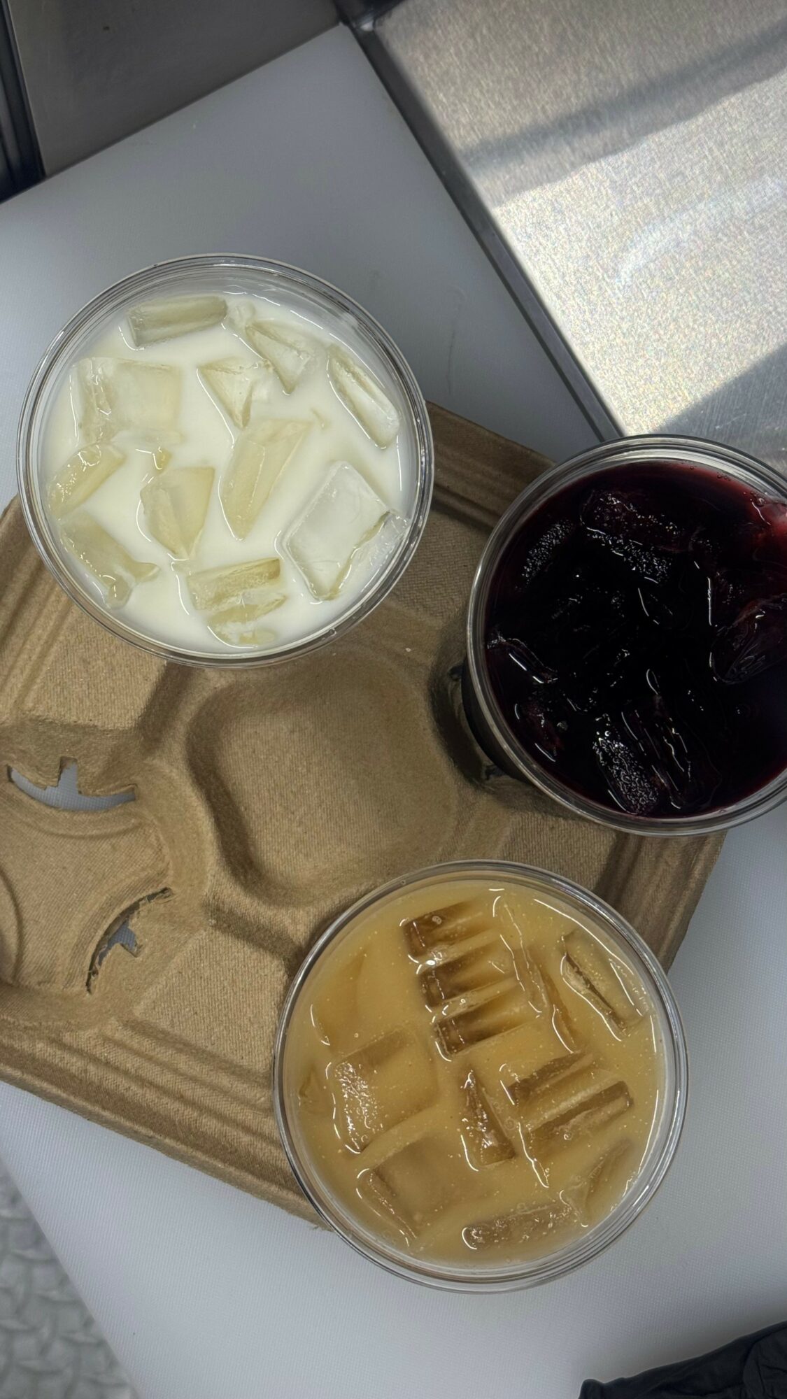 Three glasses with different beverages on a tray, one with white liquid and ice, one with dark liquid, and one with light brown liquid and ice.