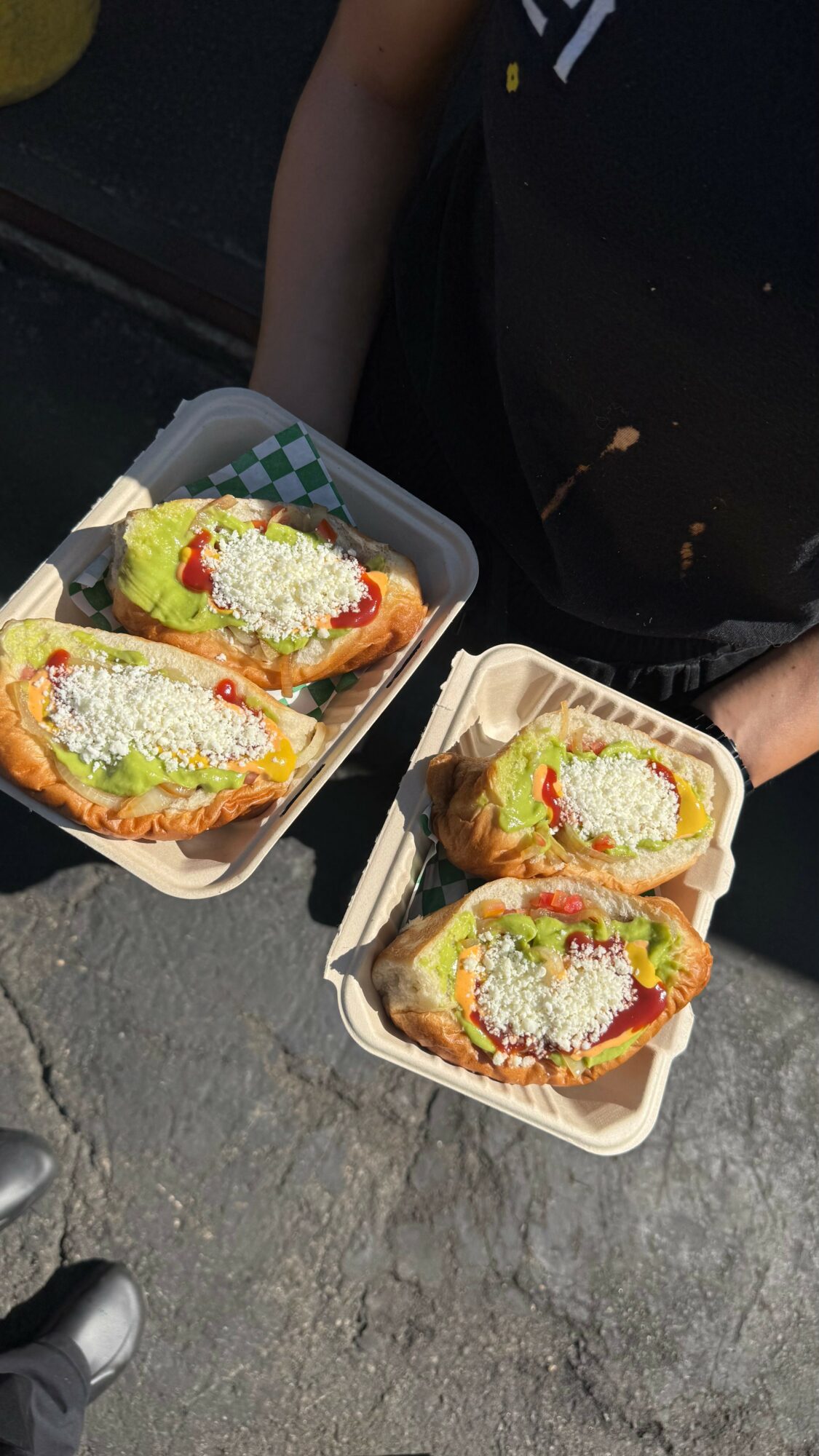 Two people holding trays with tacos topped with green sauce, cheese, and vegetables outdoors on a gray surface.