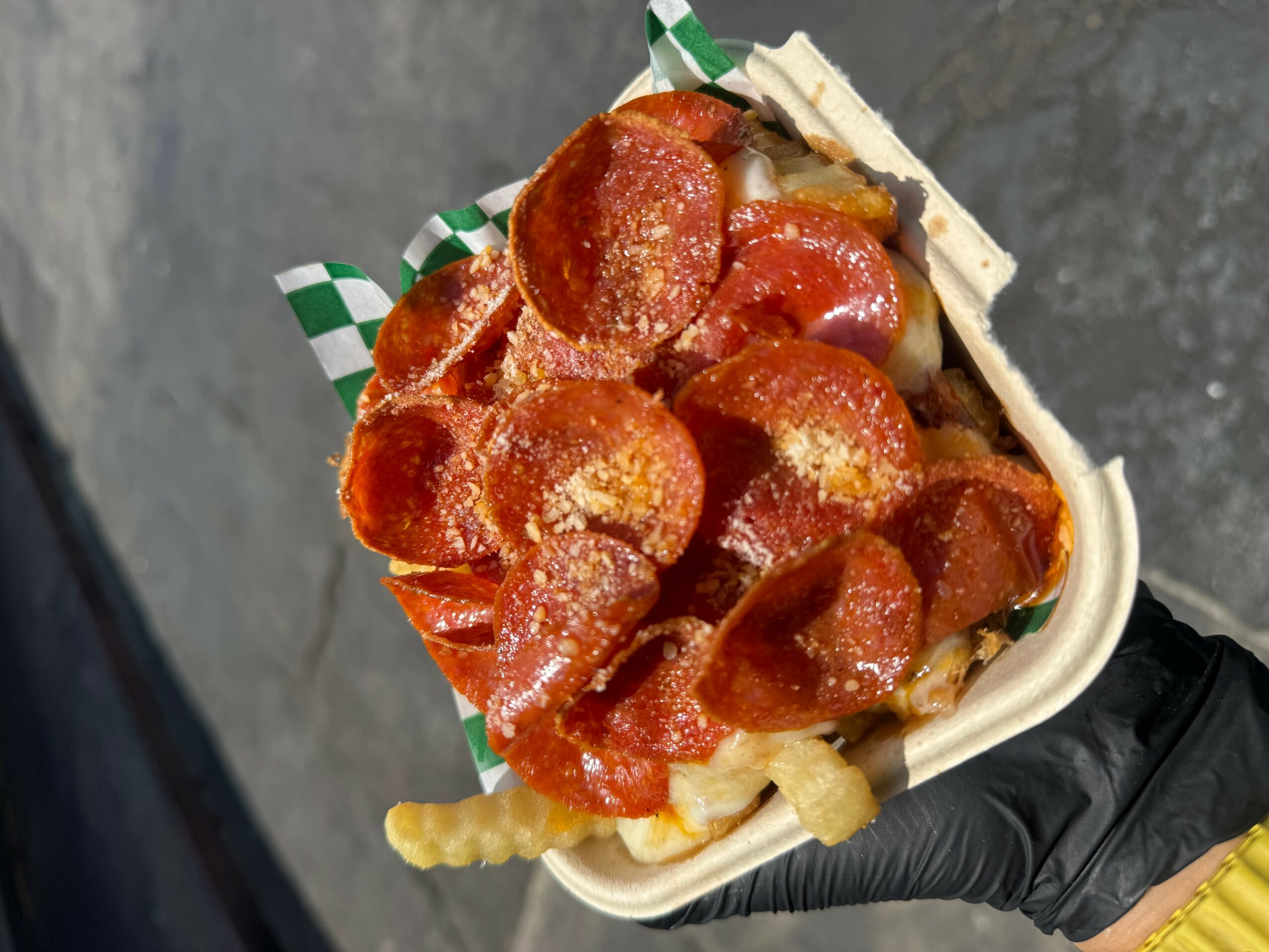 Hand holding a container of loaded fries topped with pepperoni slices and cheese, with a checkered paper liner.