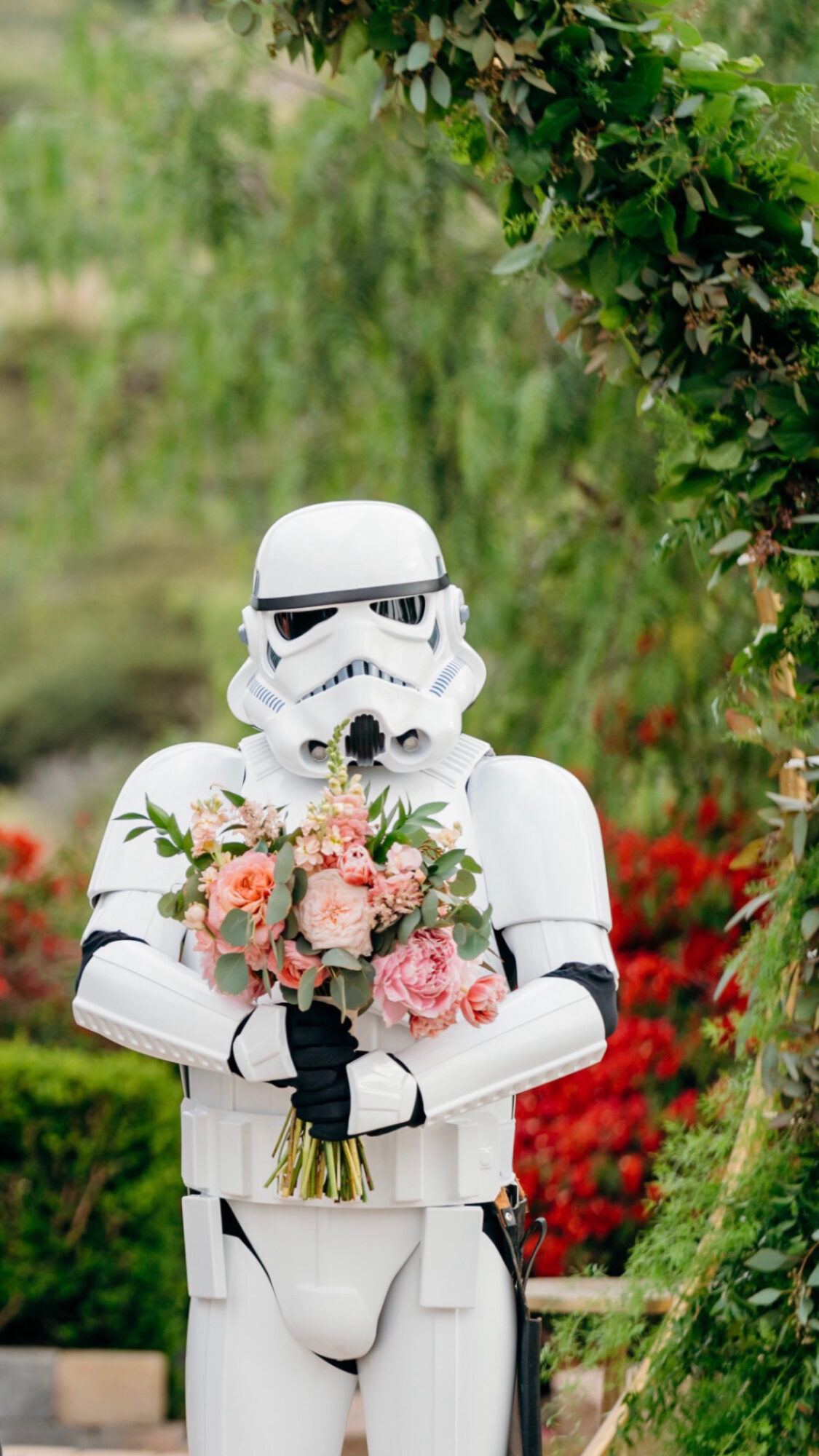 Person in Stormtrooper costume holding a bouquet of pink and white flowers outdoors.