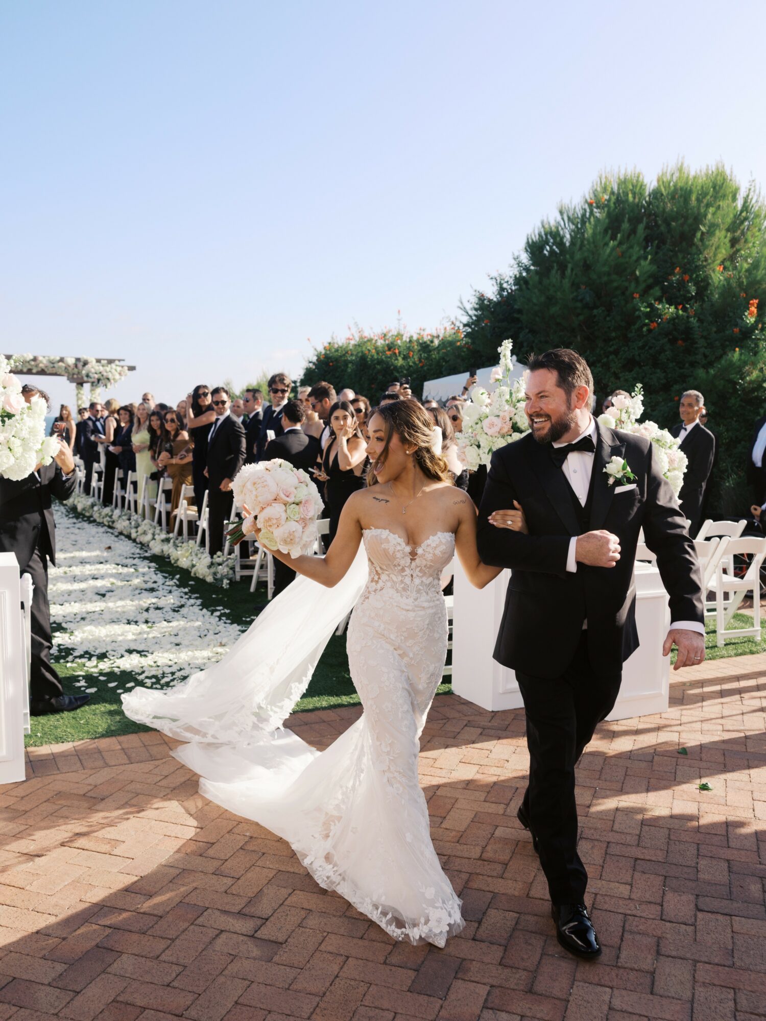 Bride and groom walking outdoors with guests and greenery in background, bride holding bouquet, groom in tuxedo.