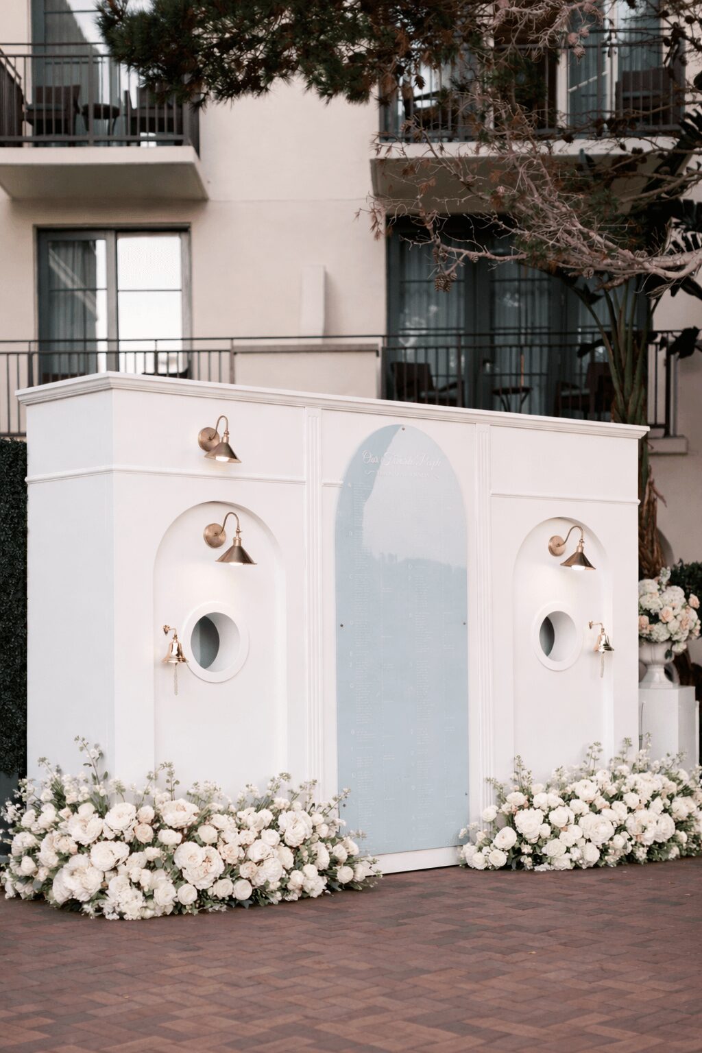 White decorative structure with floral arrangements at the base, set outdoors with a building and trees in background.