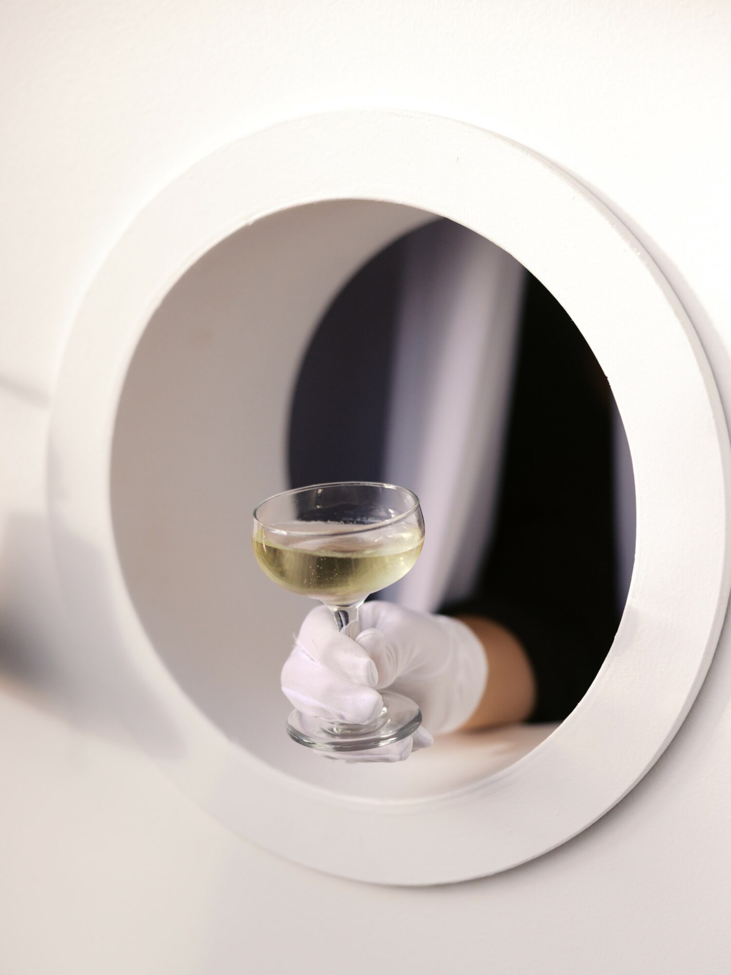 Person holding a glass of white wine through a circular opening in a white machine or device.